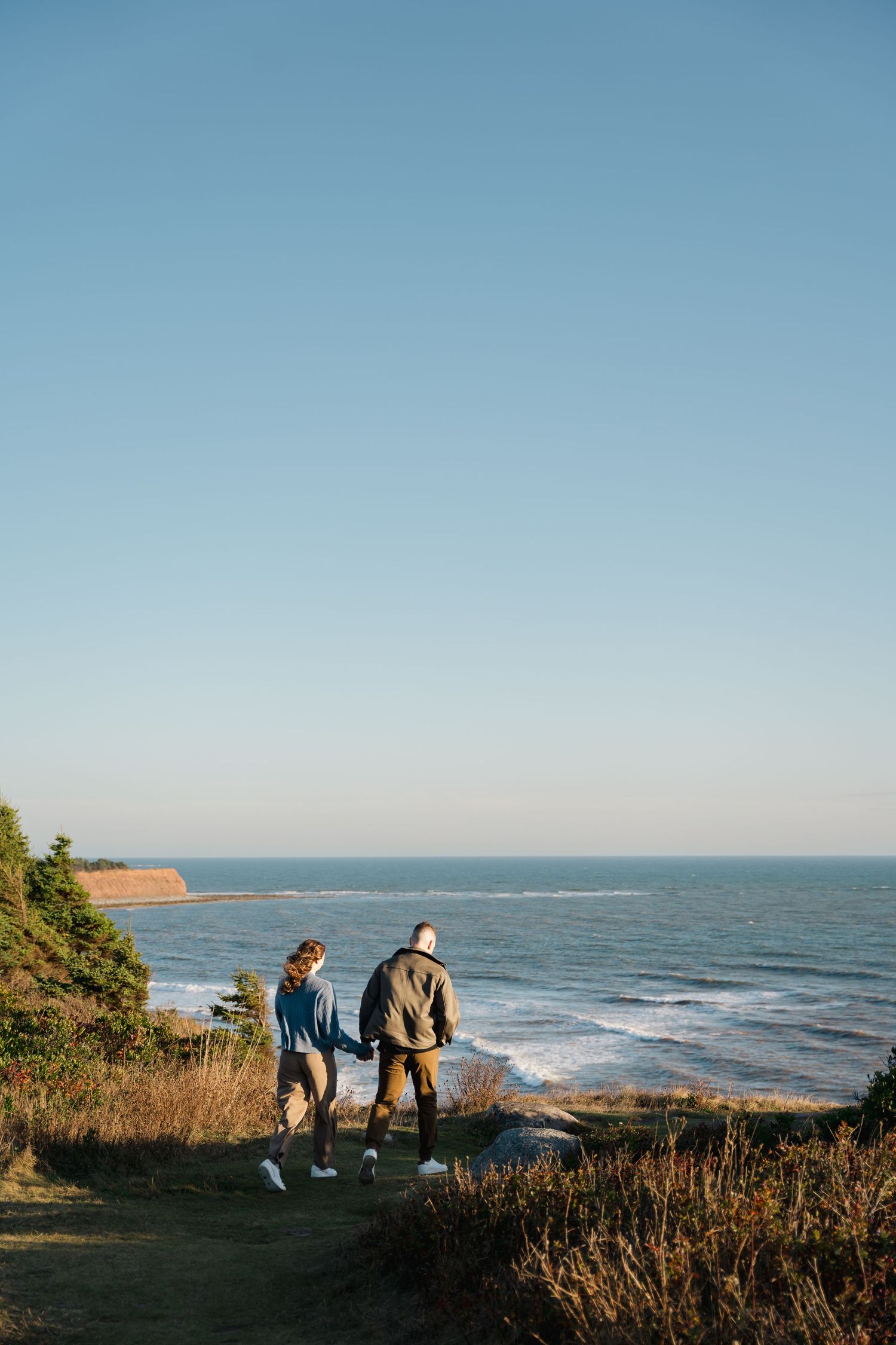 Two people walk along a coastal trail overlooking the ocean waves and cliffs on a clear sunny day in Halifax, Nova Scotia.