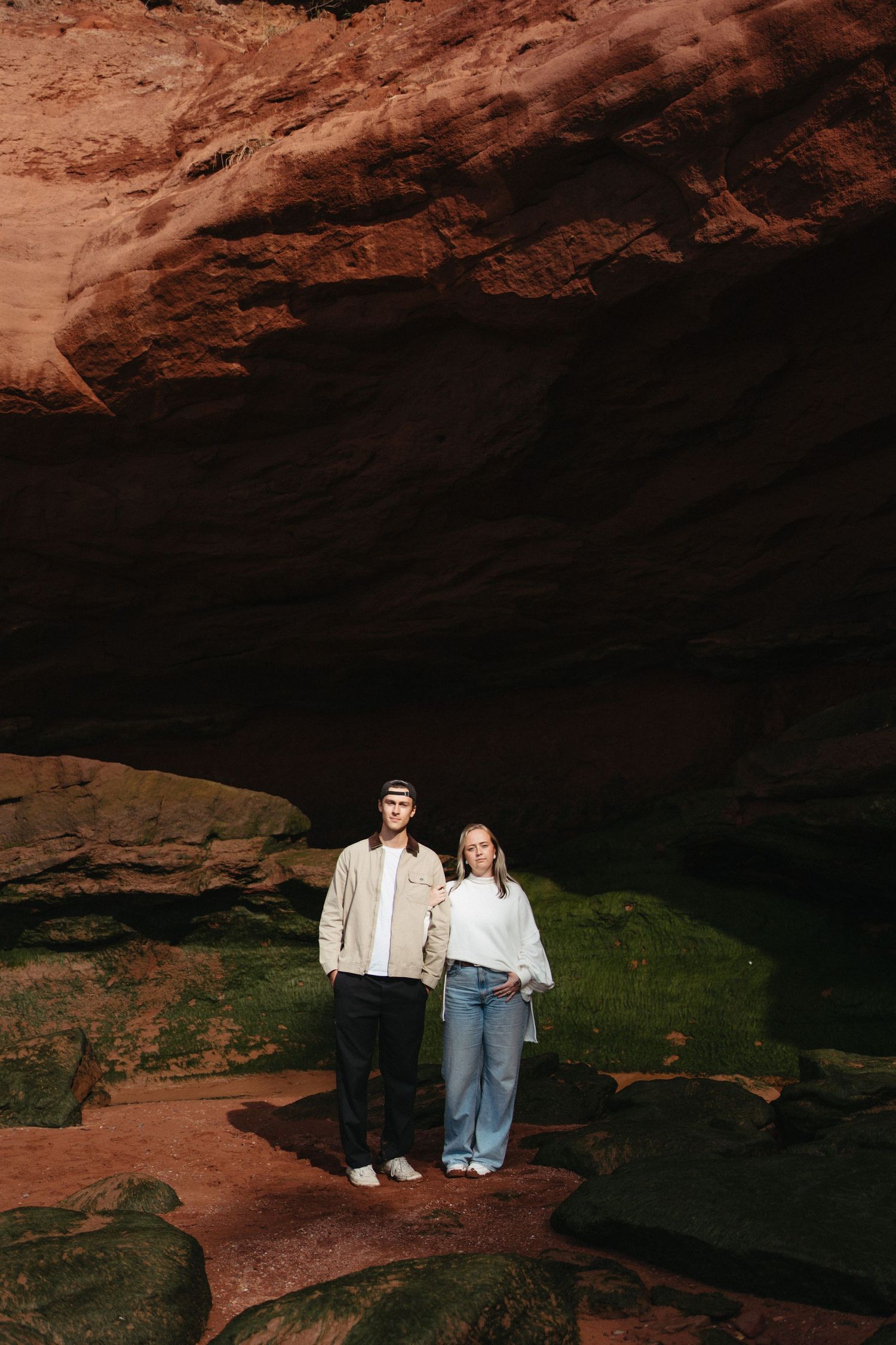 Two people stand in silhouette against red rock formations in a cave-like setting with dramatic natural lighting in Nova Scotia.