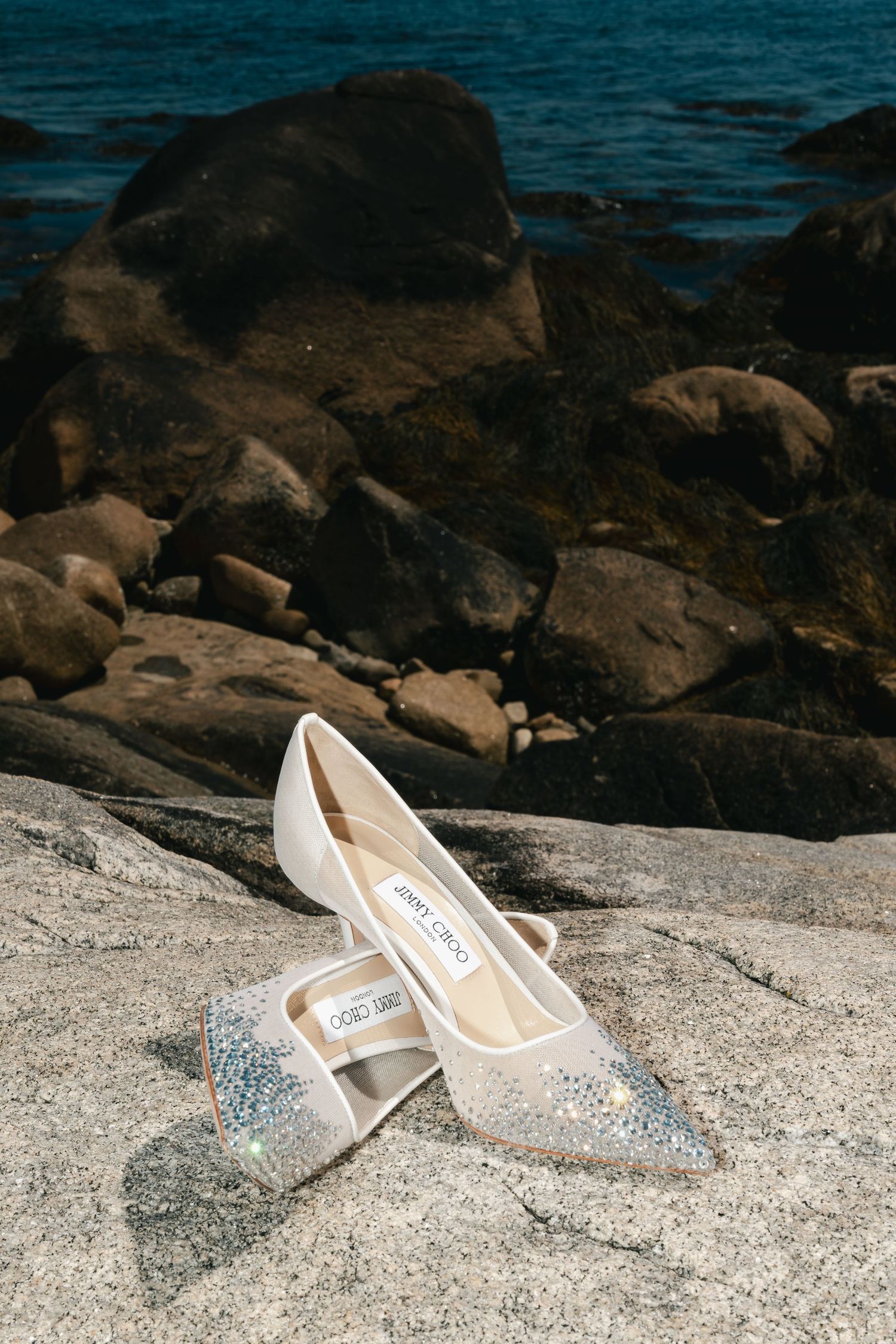 White Jimmy Choo wedding shoe with glittery details rest on a rocky beach shore with dark ocean waters in the background.