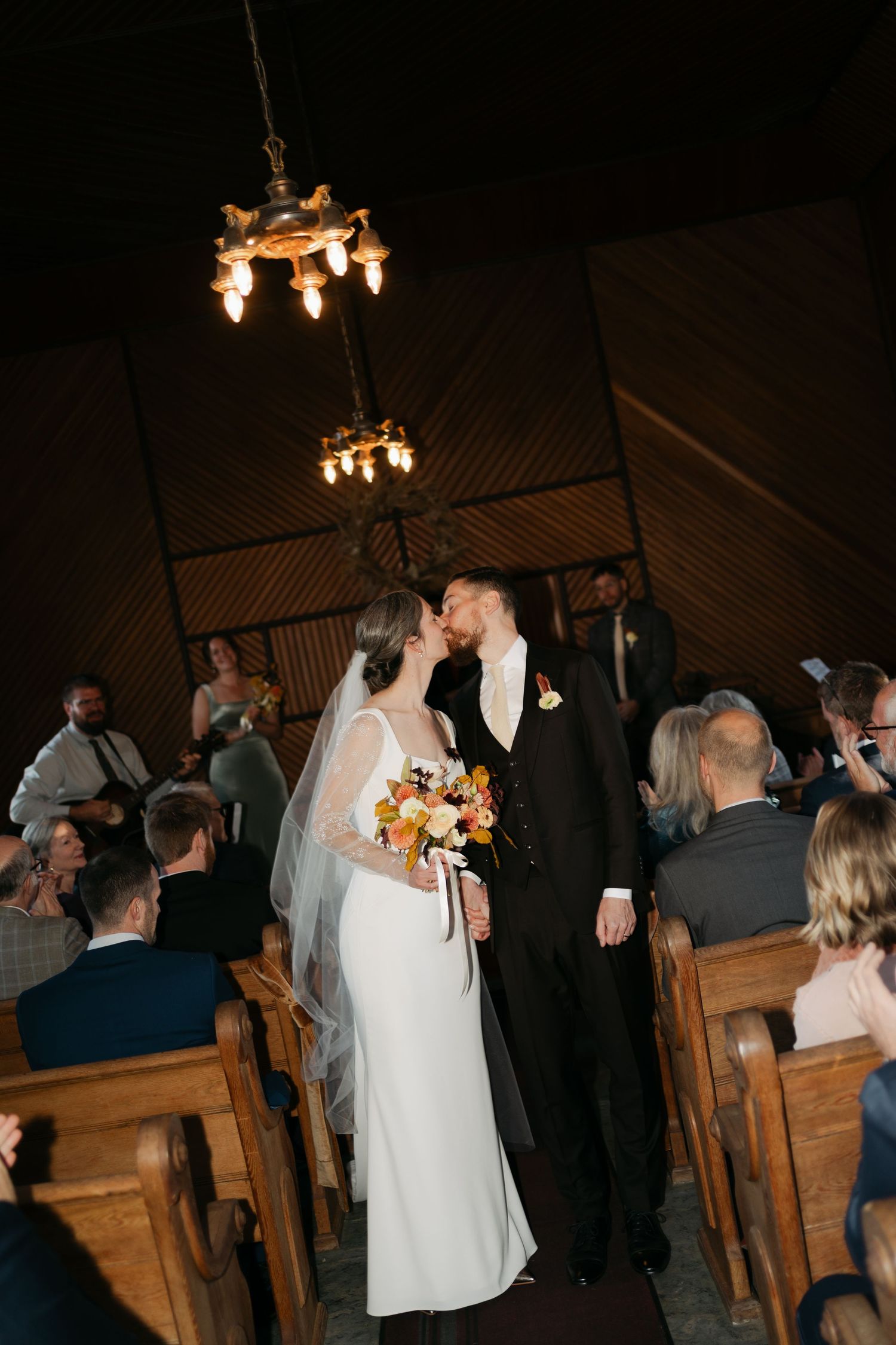 A couple shares their first kiss during a wedding ceremony in a rustic chapel with wooden pews and hanging chandeliers in Nova Scotia.