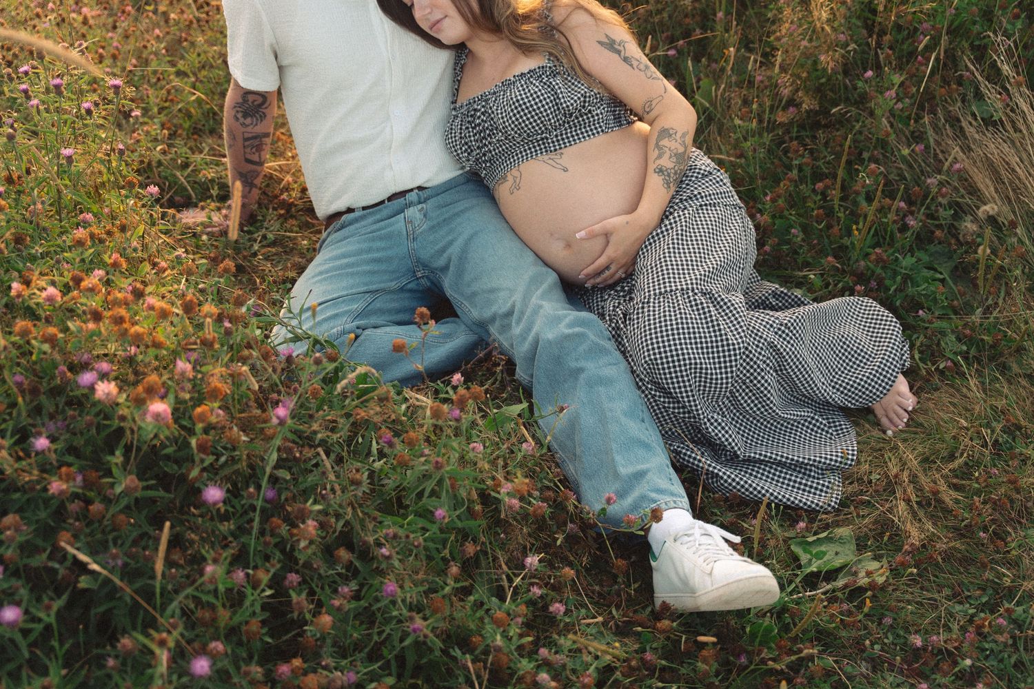 A couple sits on grass surrounded by wildflowers while wearing jeans and a white shirt and a gingham dress in Lawrencetown, Halifax.
