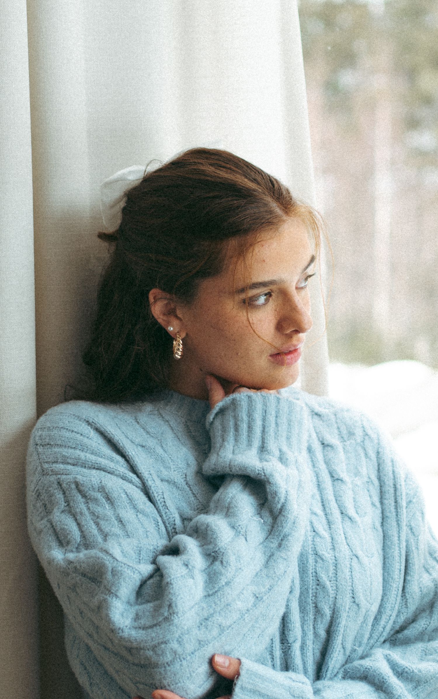 Person wearing a cozy light blue cable knit sweater gazes thoughtfully out a window in soft natural light in Halifax, Nova Scotia.