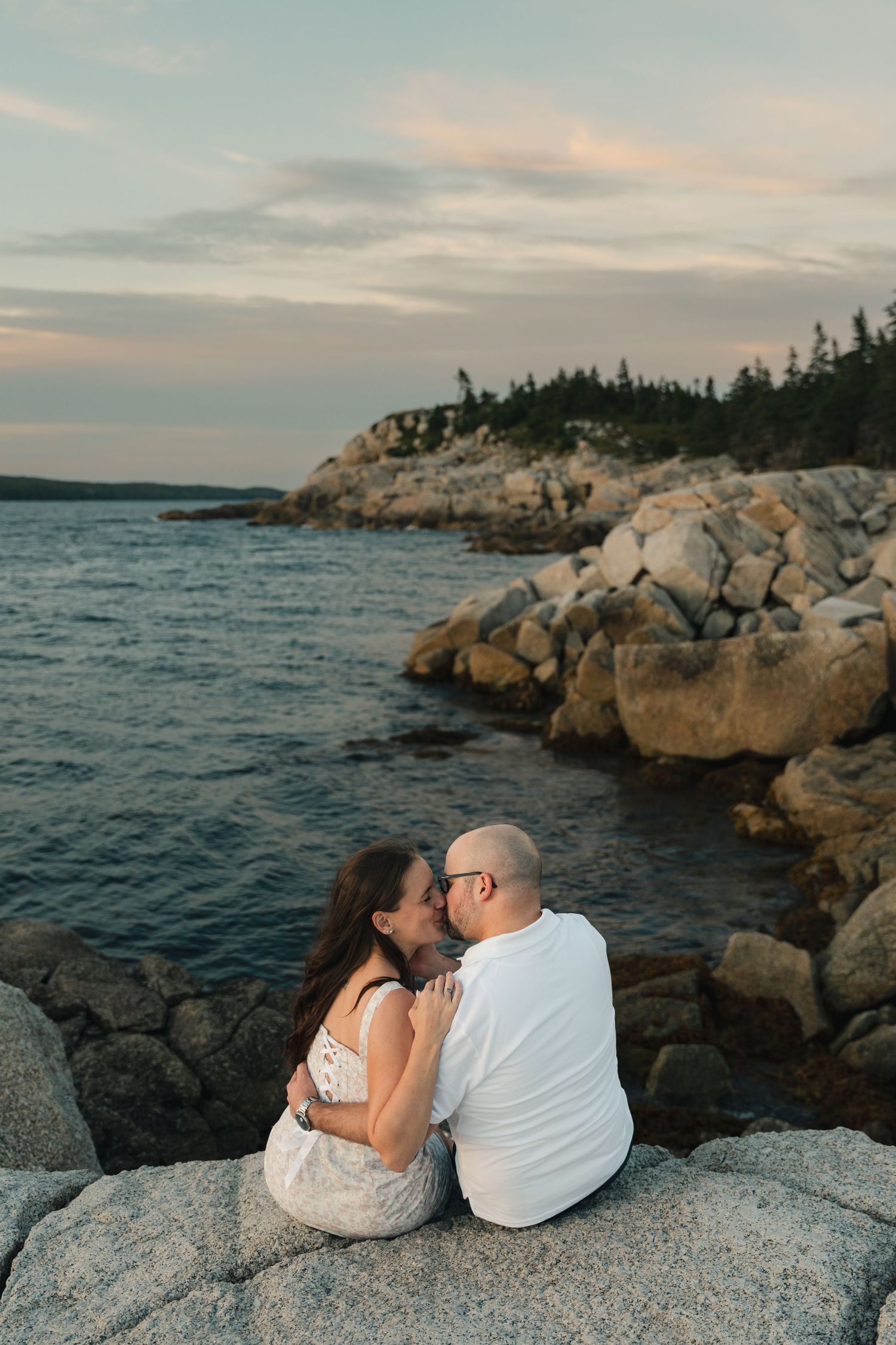 Couple sits together on rocky coastal outcrop at sunset overlooking the ocean in Nova Scotia.