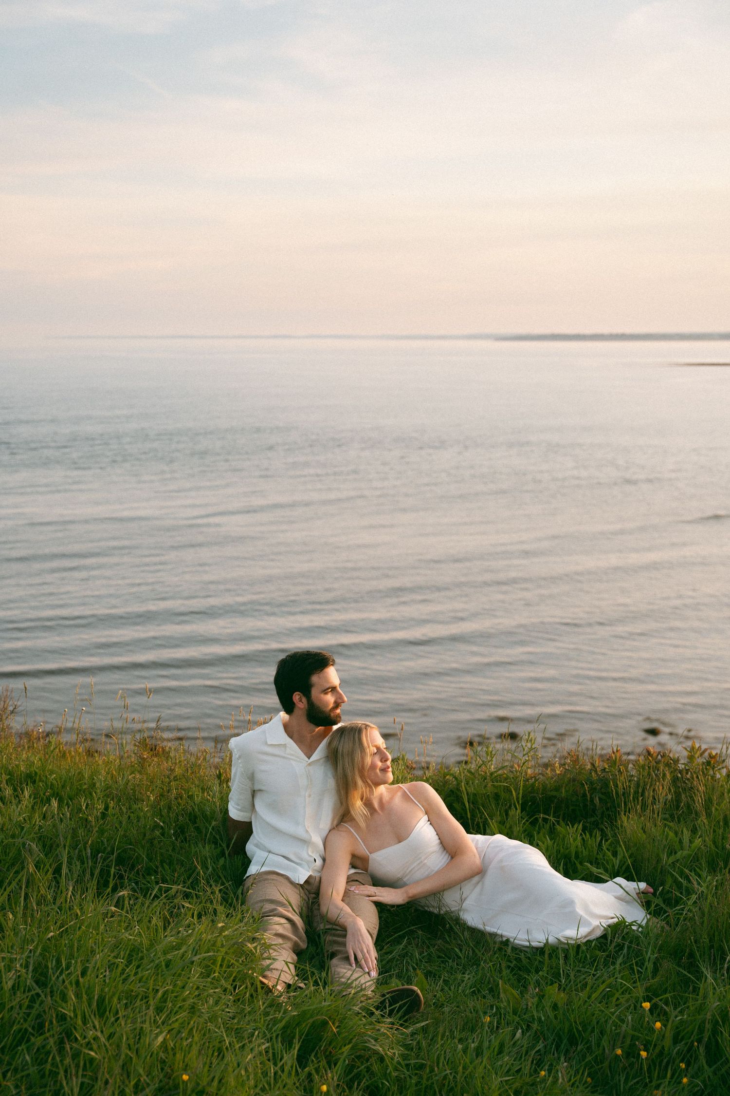 A couple in wedding attire relaxes on grassy cliffside  in Halifax, Nova Scotia overlooking a serene ocean view at sunset.