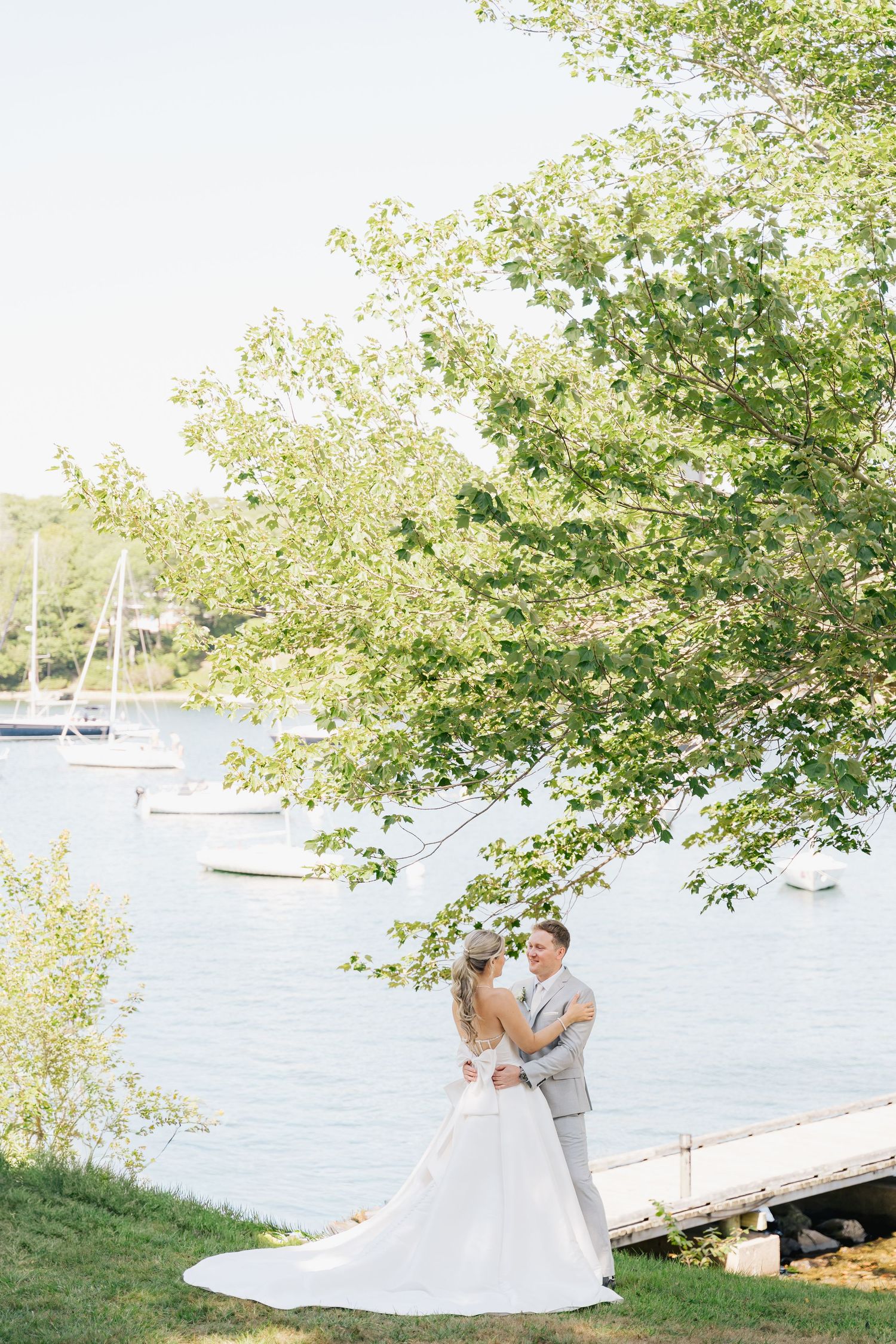 A couple in wedding attire dances together on a wooden dock by a scenic waterfront with boats and trees in view in Halifax, Nova Scotia.
