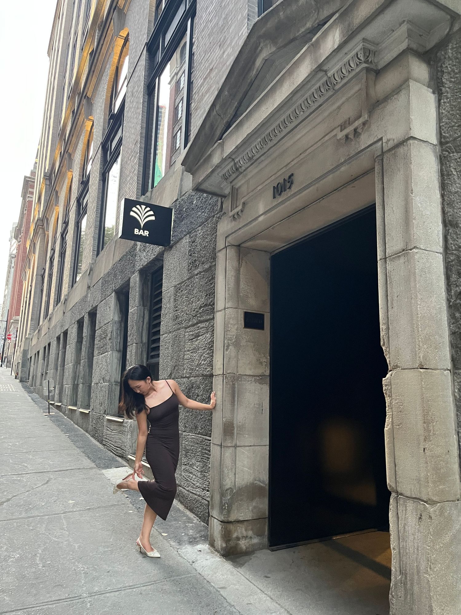 A person in a black dress poses elegantly against a stone building entrance with a flag hanging above.