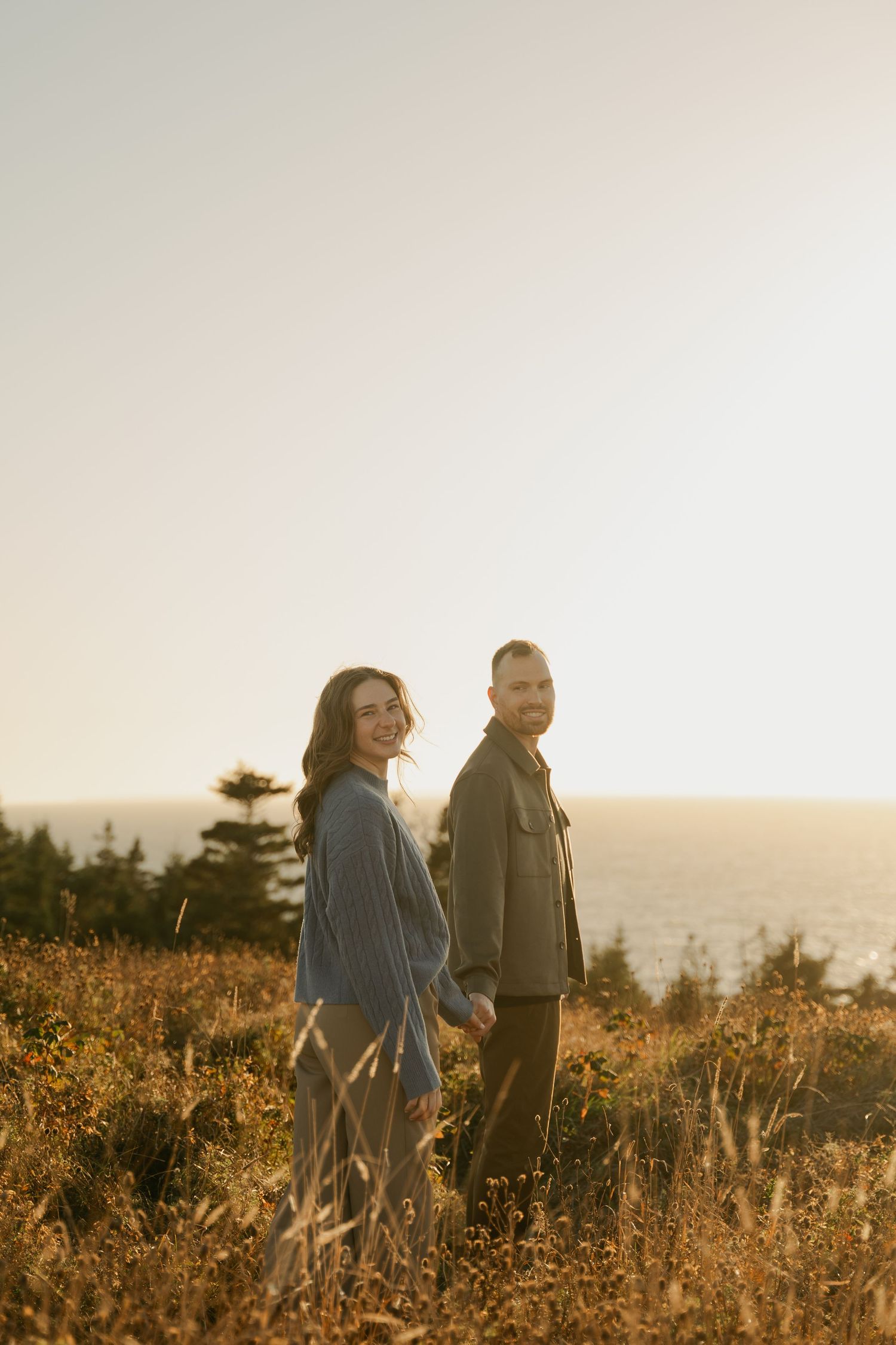 A couple walks together through a golden meadow at sunset with the ocean visible in the distance in Halifax, Nova Scotia.