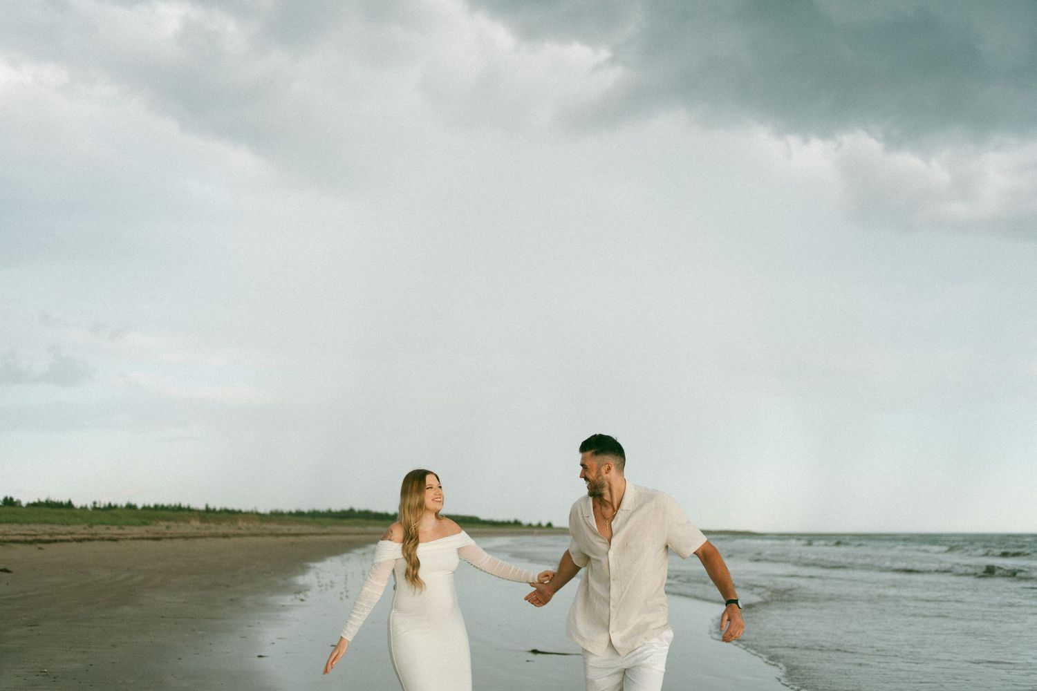 A couple in white attire runs playfully along a beach with stormy clouds overhead and waves crashing nearby in Halifax, Nova Scotia.