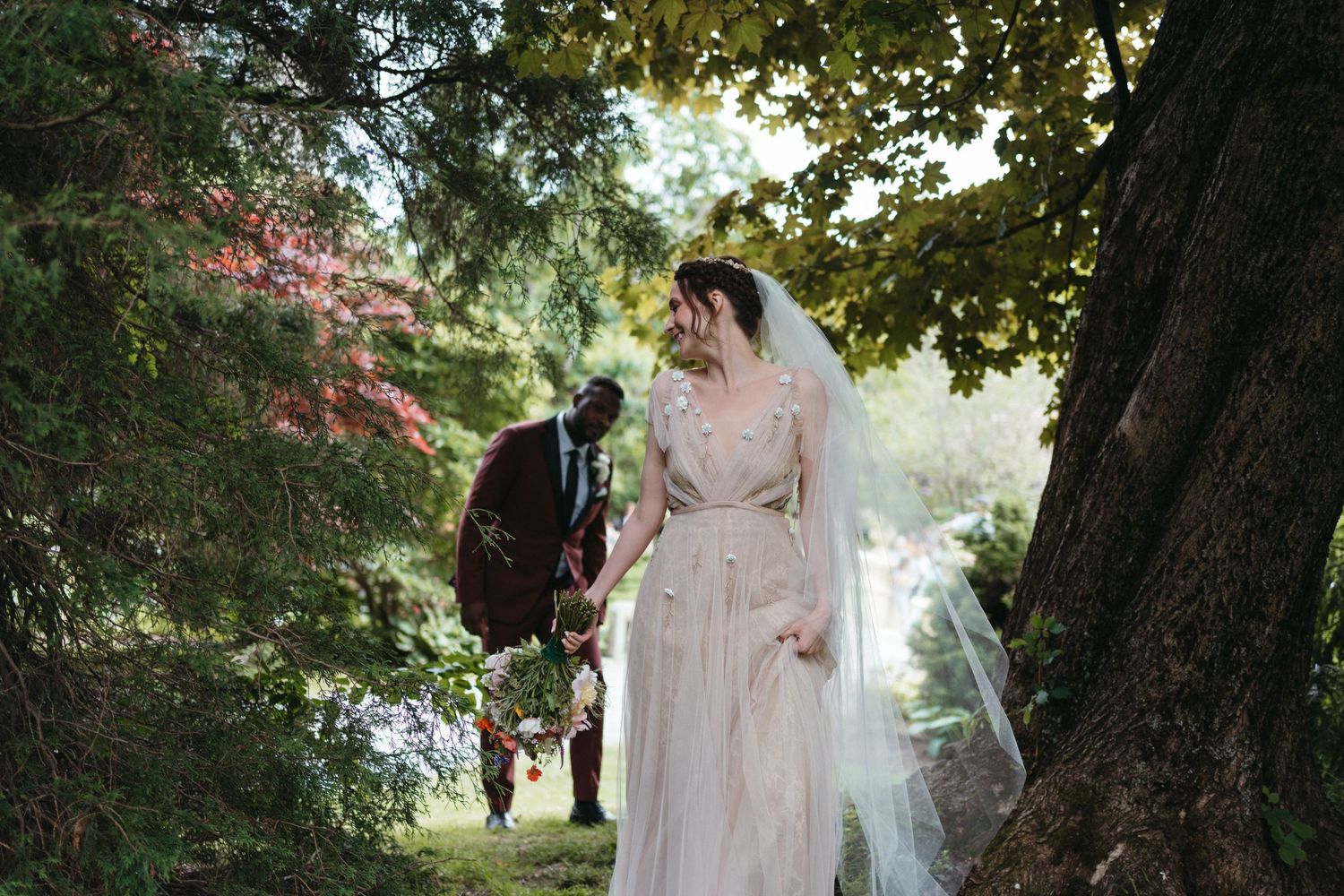 A couple in wedding attire walks through a lush garden path with sunlight filtering through the trees in Halifax, Nova Scotia.