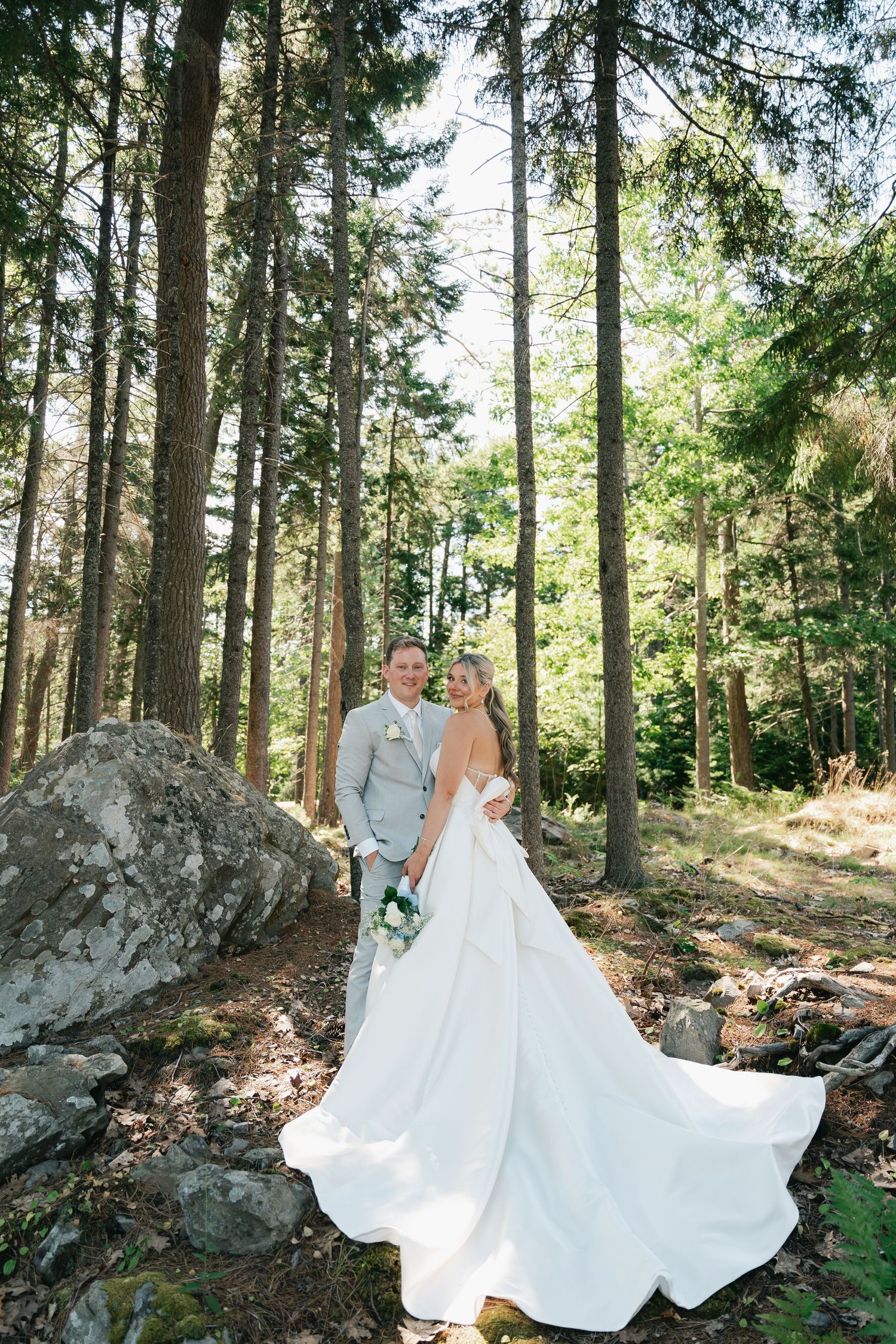 Wedding couple poses in sunlit forest with bride's dress flowing dramatically among tall trees in Halifax, Nova Scotia.