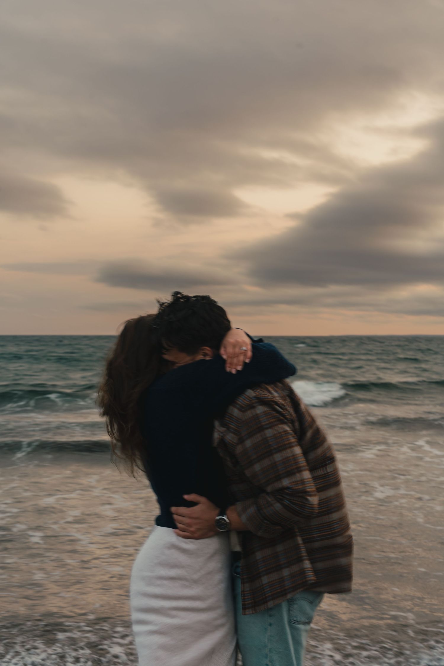 Two people share an intimate moment on a beach in Halifax, Nova Scotia at sunset with dramatic waves and cloudy skies.