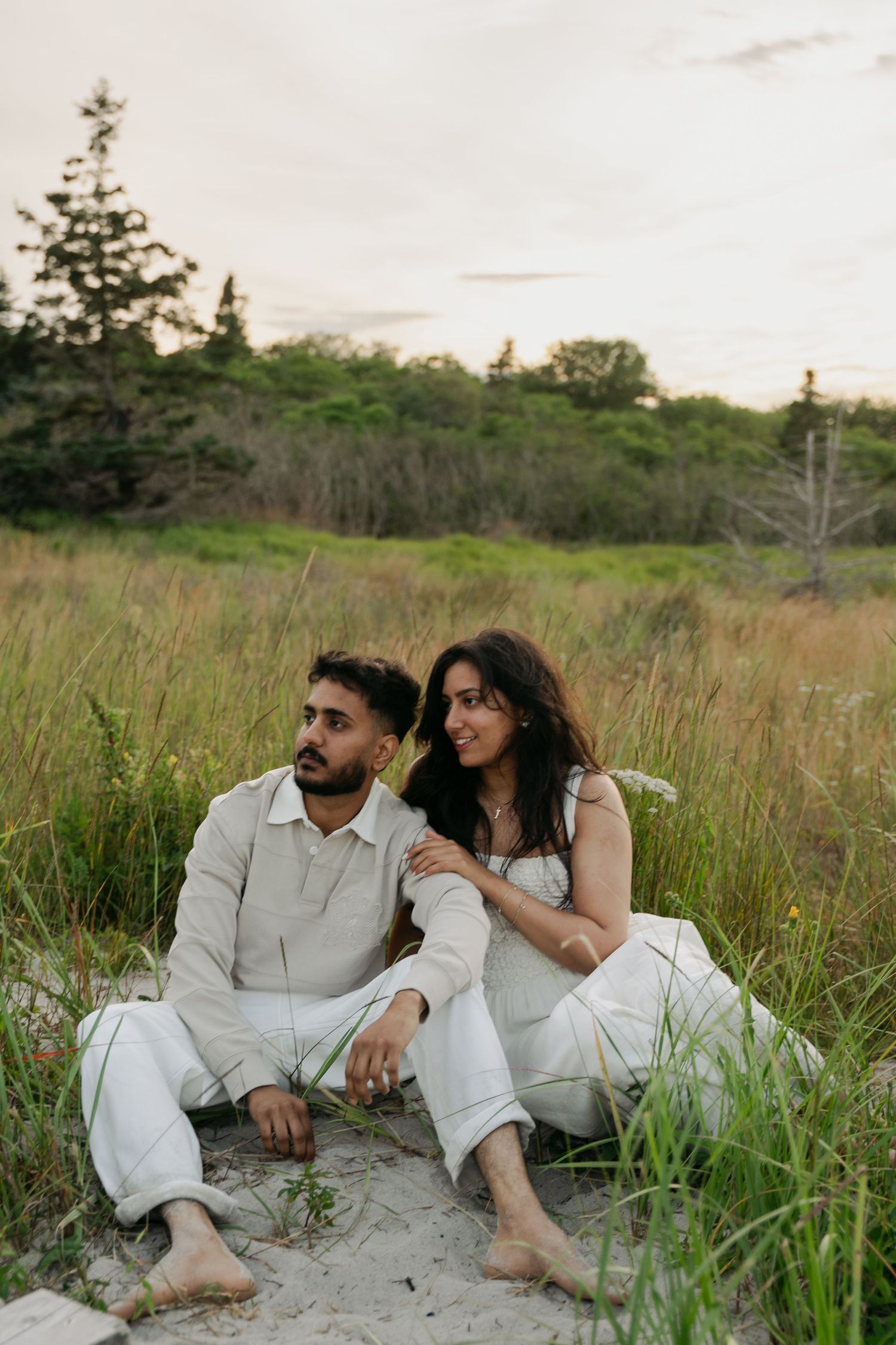 Two people in white clothing sit together on sandy ground surrounded by tall grass in a natural outdoor setting in Halifax, Nova Scotia.