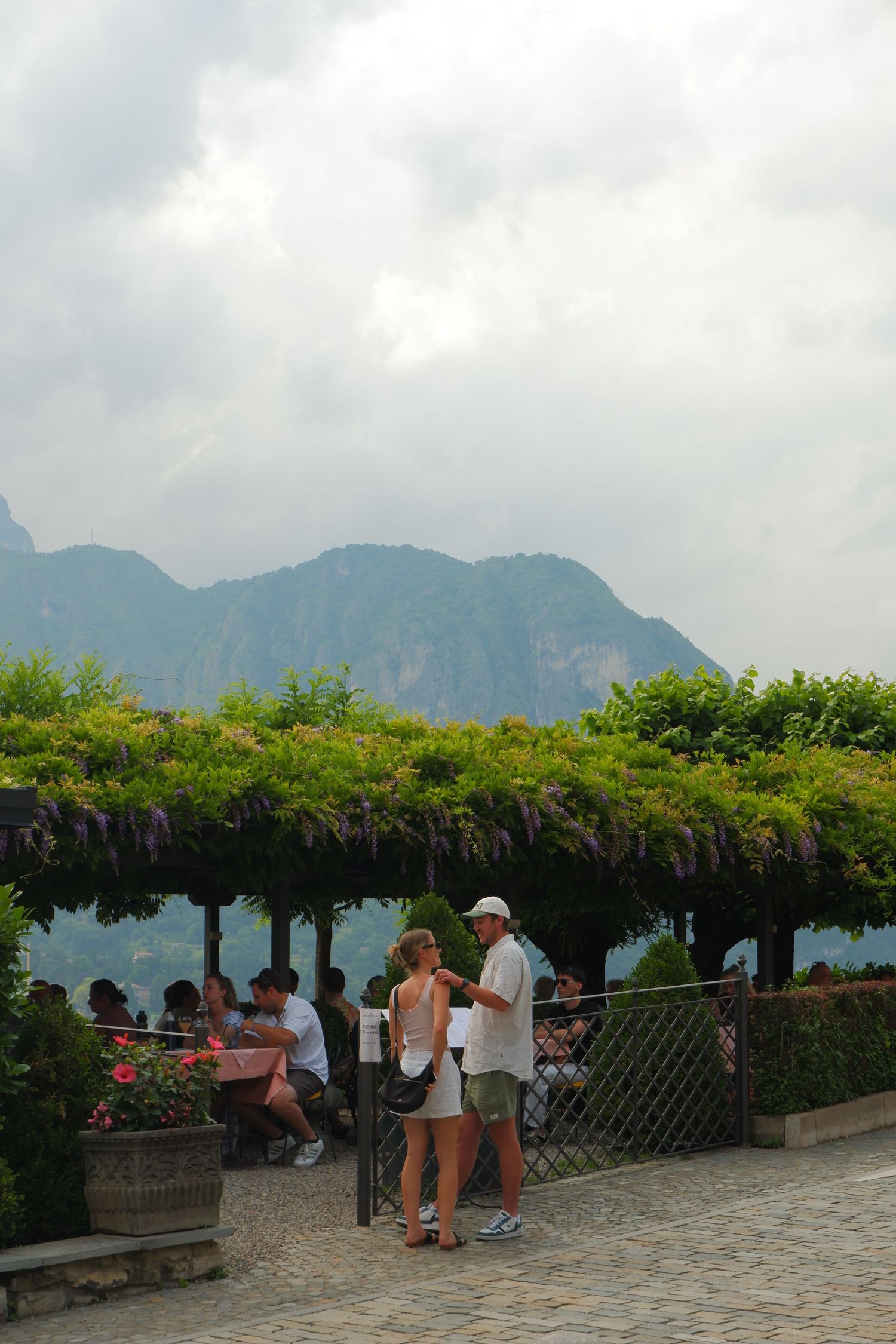 Tourists enjoy a scenic mountain view from a wisteria-covered pergola terrace in Lake Como, Italy.