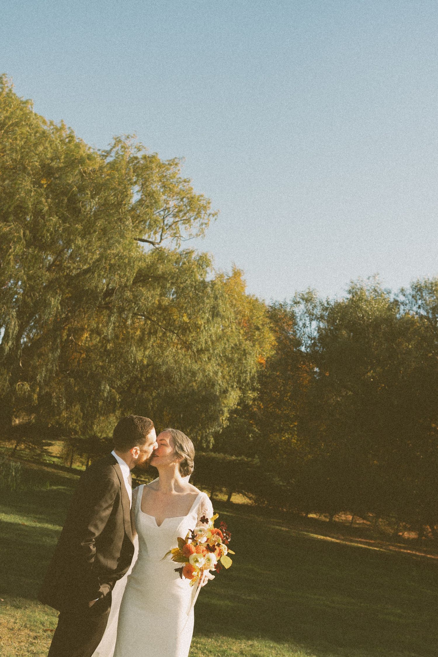 Wedding couple stands together in golden afternoon sunlight with autumn trees in background in Nova Scotia.