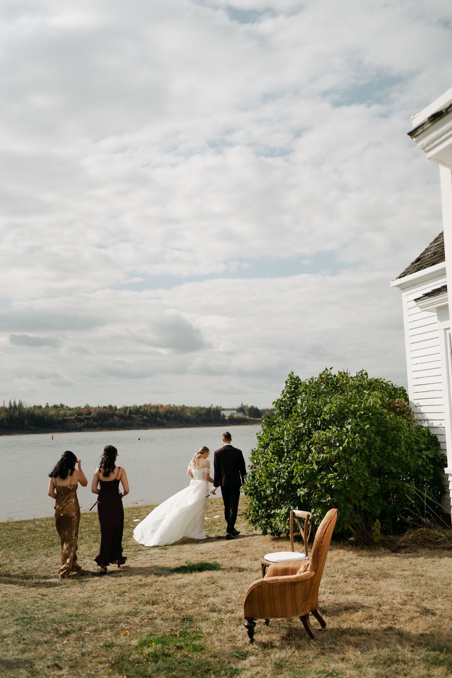 Wedding party walks along a scenic waterfront path with cloudy skies and greenery in Nova Scotia.