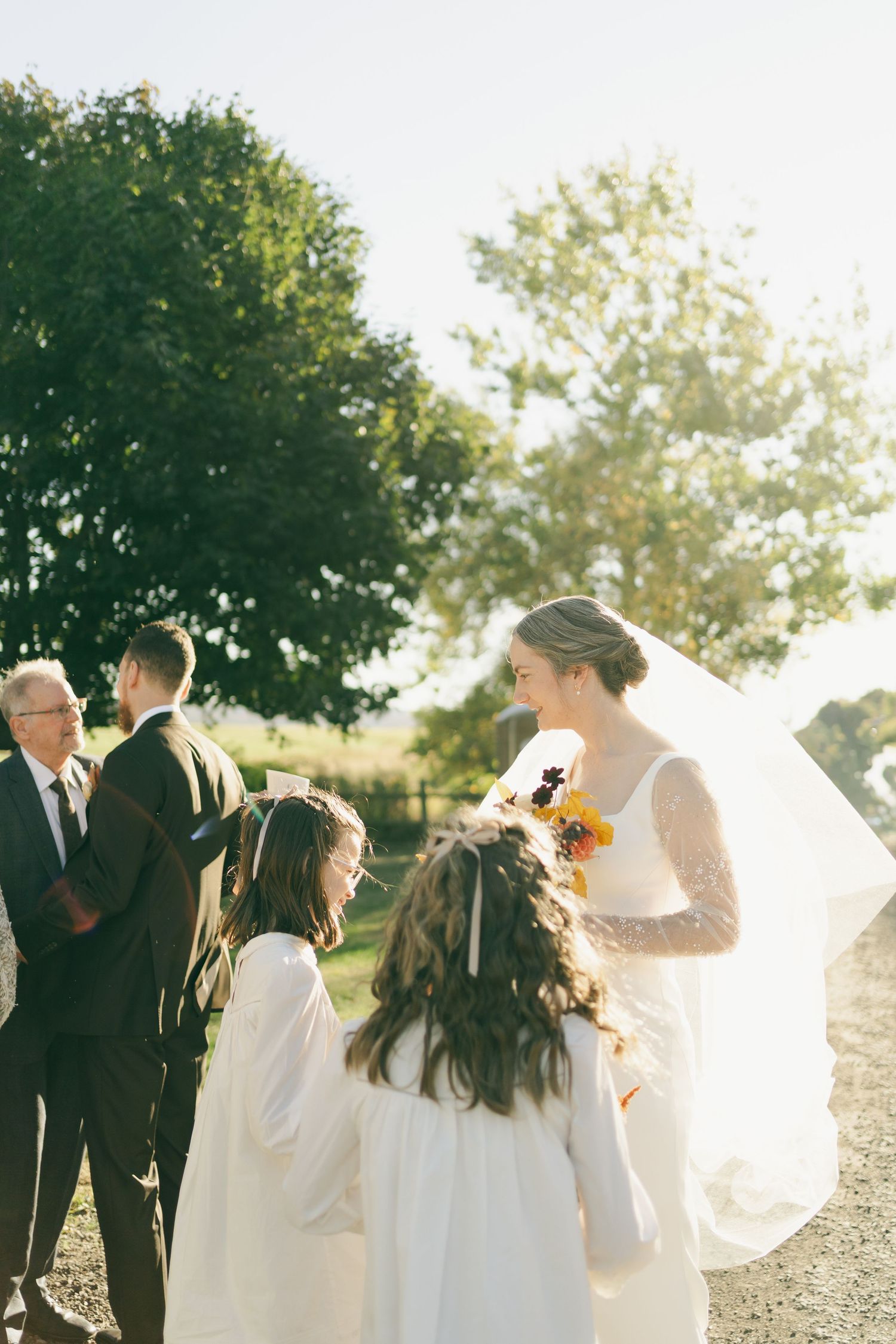 A wedding scene outdoors with flower girls in white dresses and a flowing veil catching sunlight through the trees in Nova Scotia.