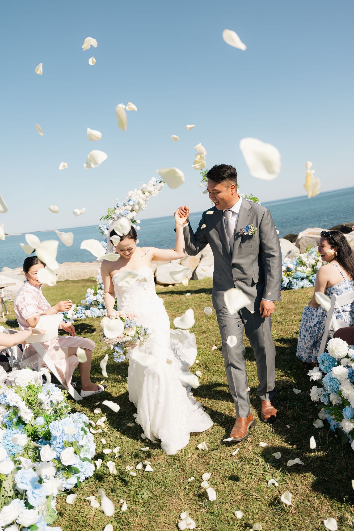 Wedding party members playfully toss white flower petals in the air on a grassy oceanside cliff during a celebration in Halifax, Nova Scoti.