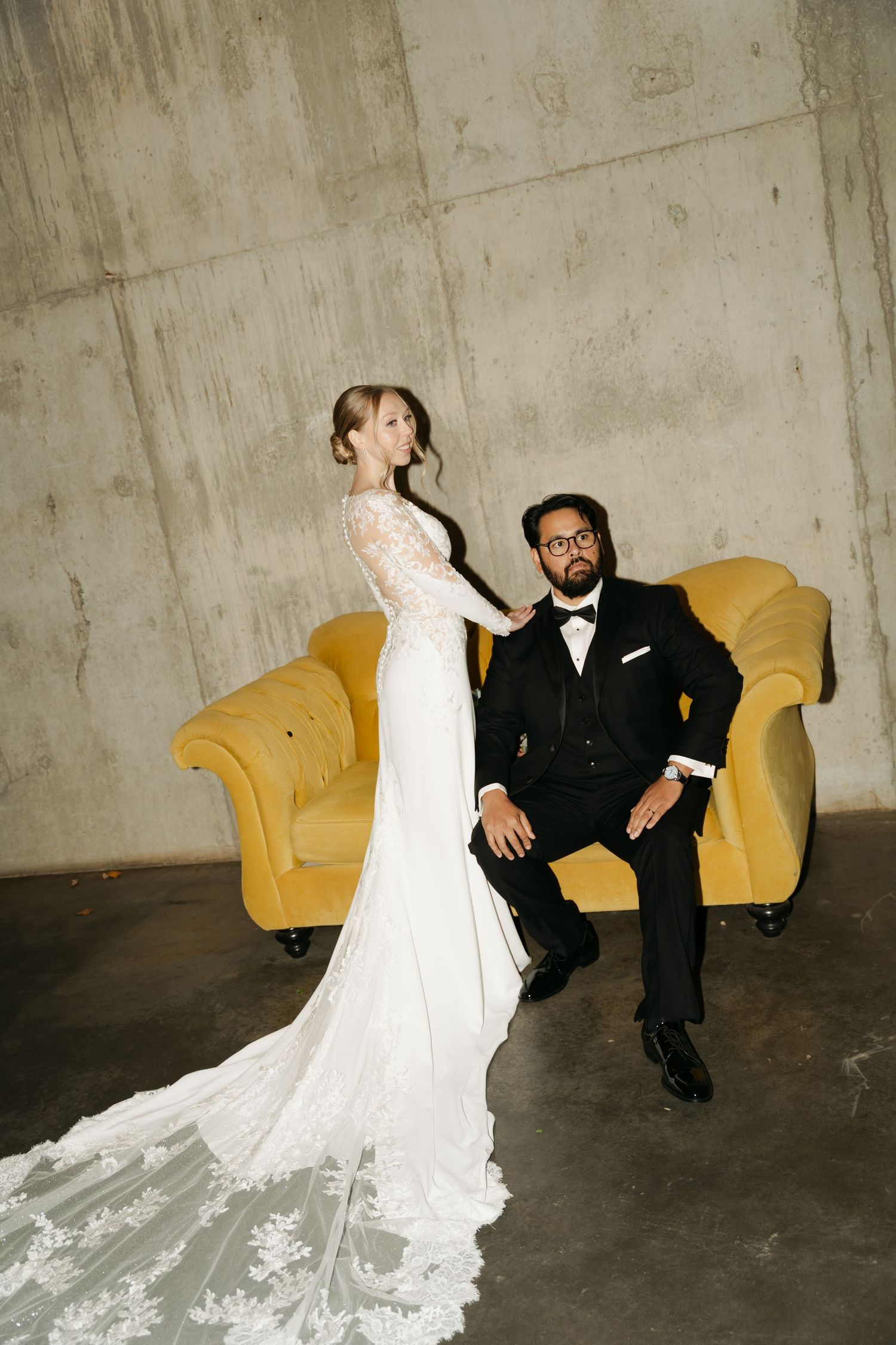 A couple in wedding attire poses playfully on a yellow vintage couch against a concrete wall in a photo at Lightfoot & Wolfville.