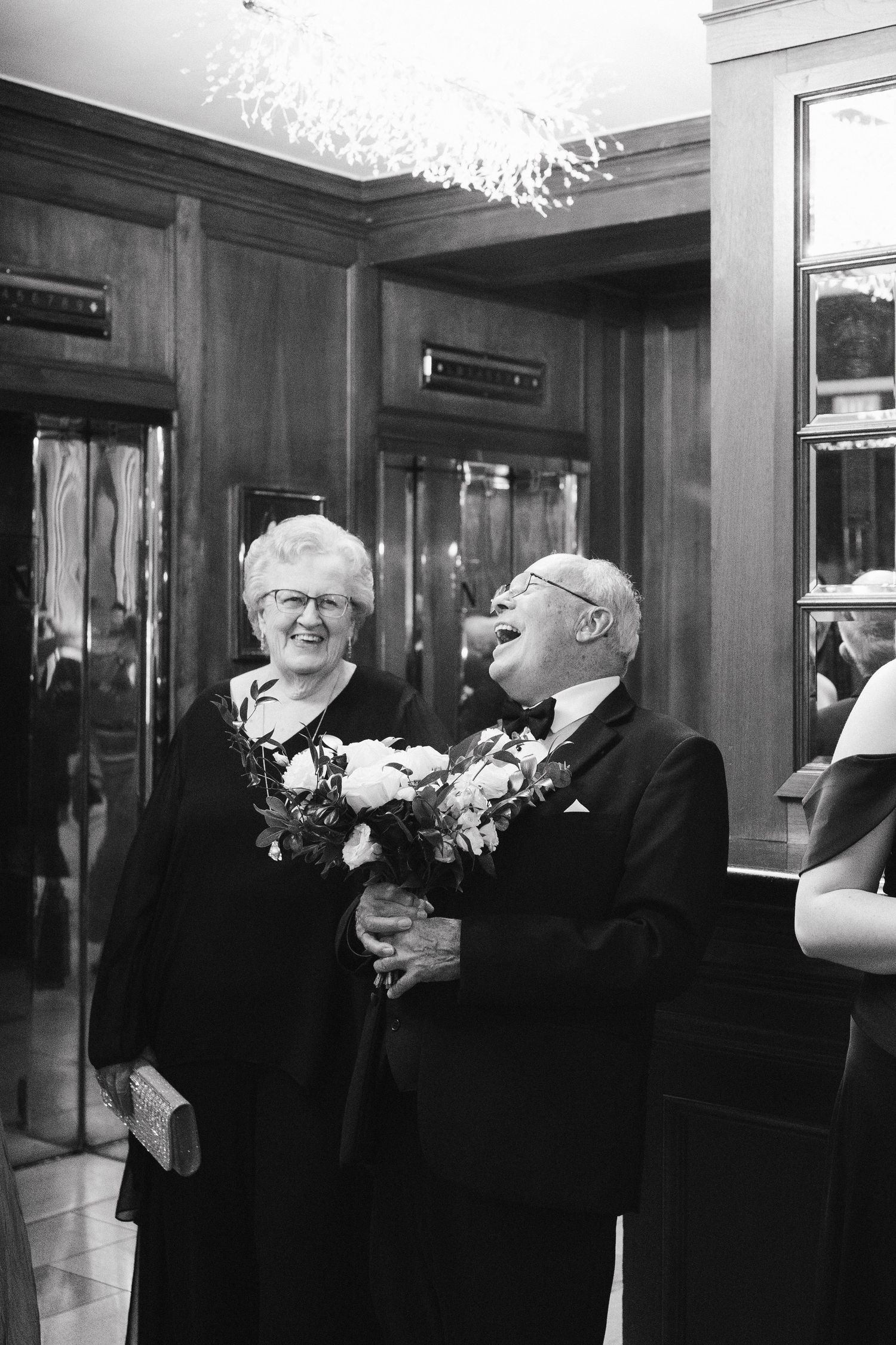 An elderly couple shares a candid joyful moment while holding bouquets of flowers in a wood-paneled hallway in Halifax, Nova Scotia.