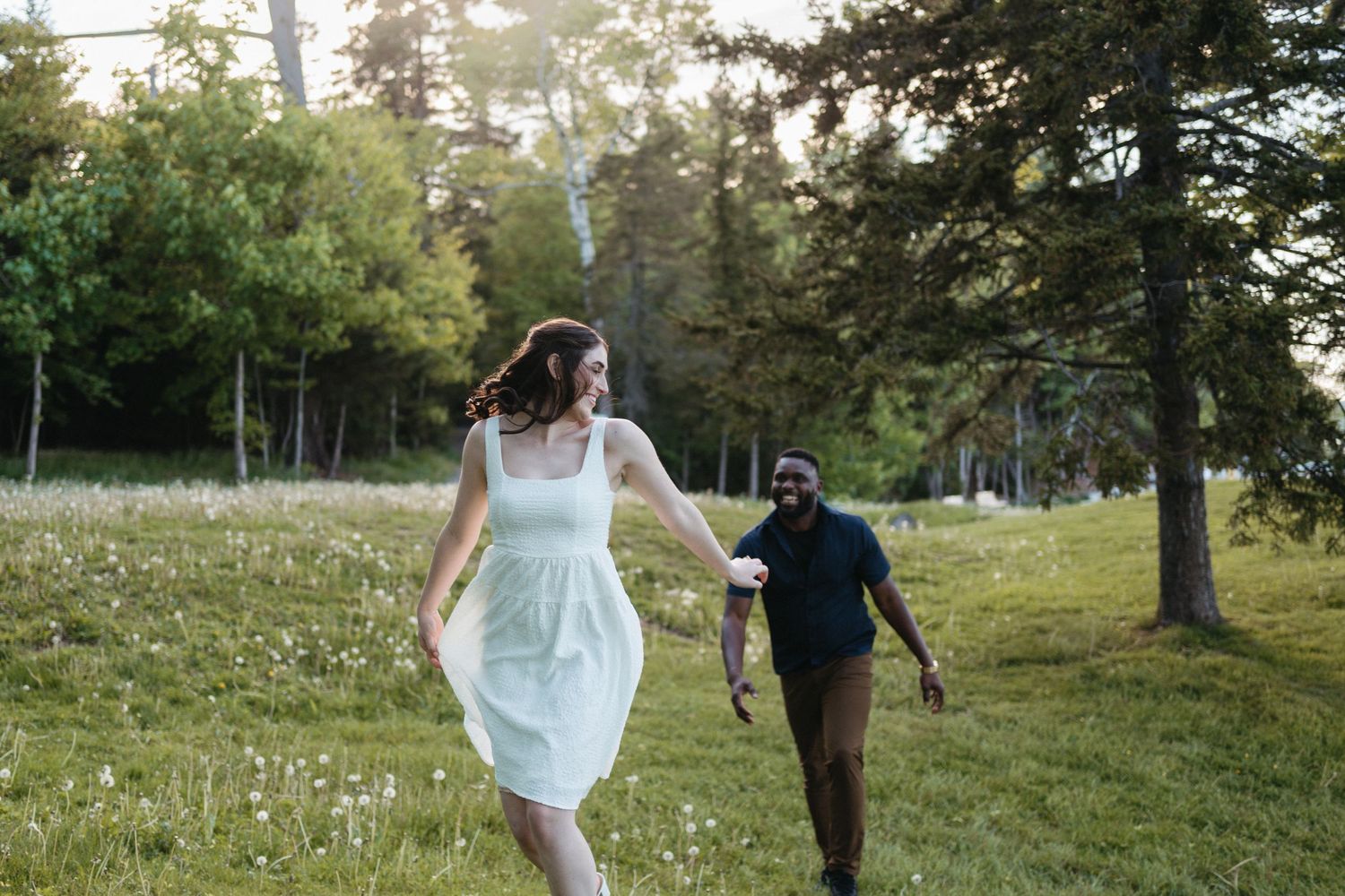 A couple walks through a lush green field at sunset with trees in the background in Halifax, Nova Scotia.