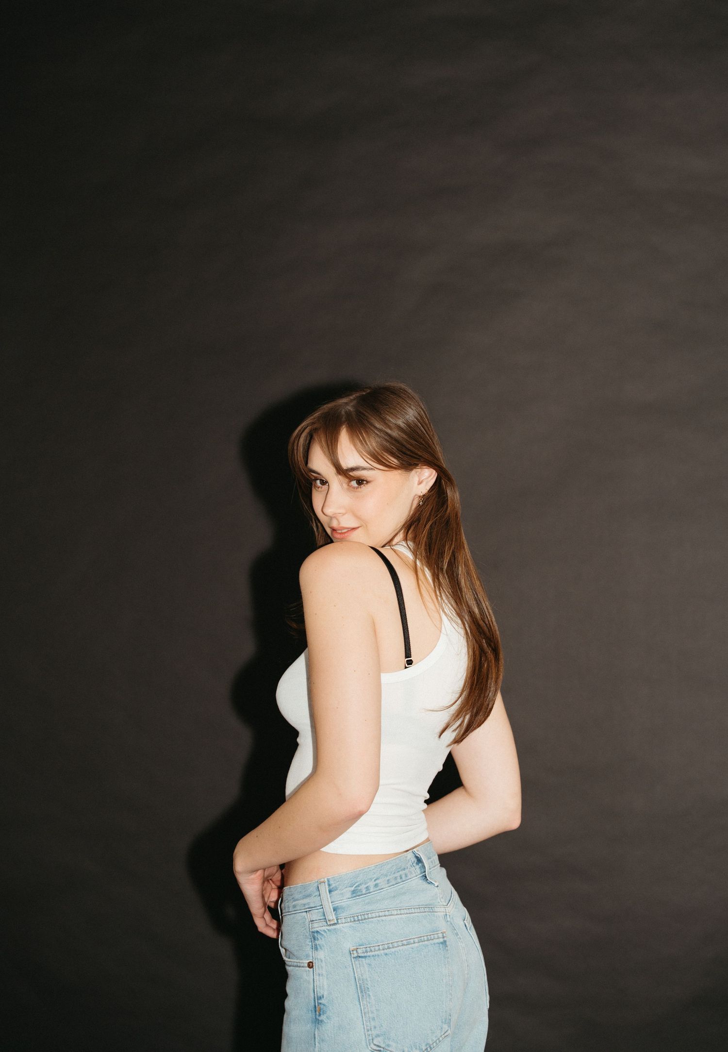 Side view portrait against dark background showing figure in white tank top and light denim jeans looking over shoulder in Halifax, NS