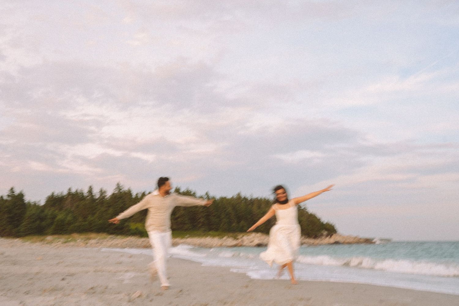 Two people in white outfits dance joyfully along a peaceful beach at sunset with trees in the background in Halifax, Nova Scotia.