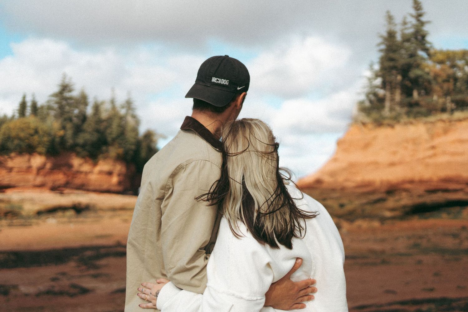 A couple stands together on a scenic mountain trail surrounded by evergreen trees and dramatic landscapes in Nova Scotia.