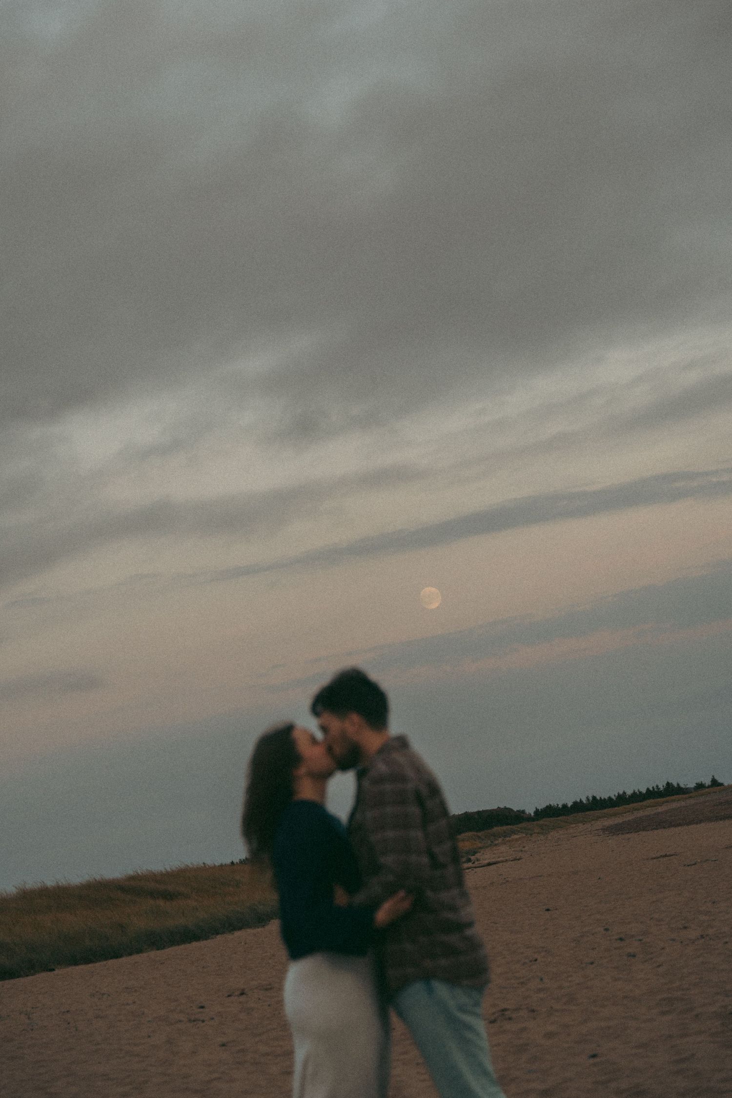 A couple embraces on a sandy beach in Halifax, Nova Scotia at dusk with moody cloudy skies and a faint moon in the background.