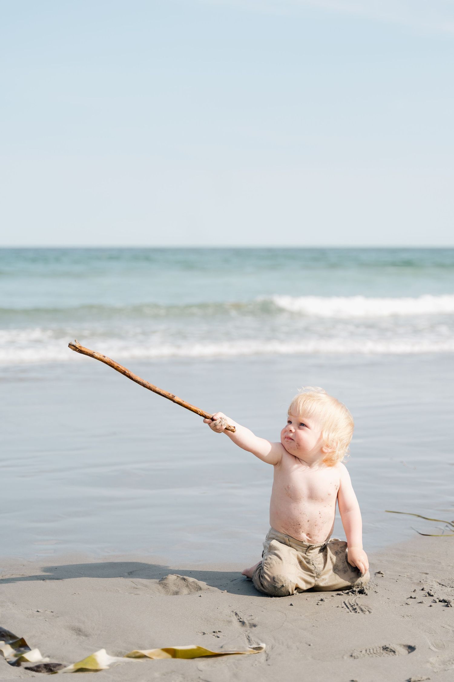 A toddler sits on a sandy beach holding a long stick while waves crash in the background in Halifax, Nova Scotia.