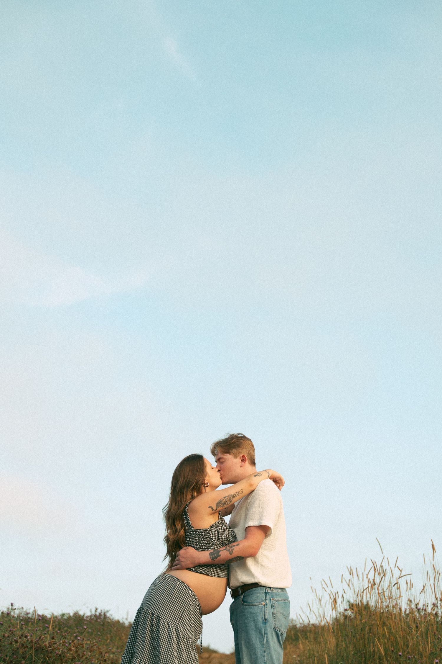 A romantic couple embraces in a hilltop meadow against a soft blue sky in Lawrencetown, Halifax, Nova Scotia.