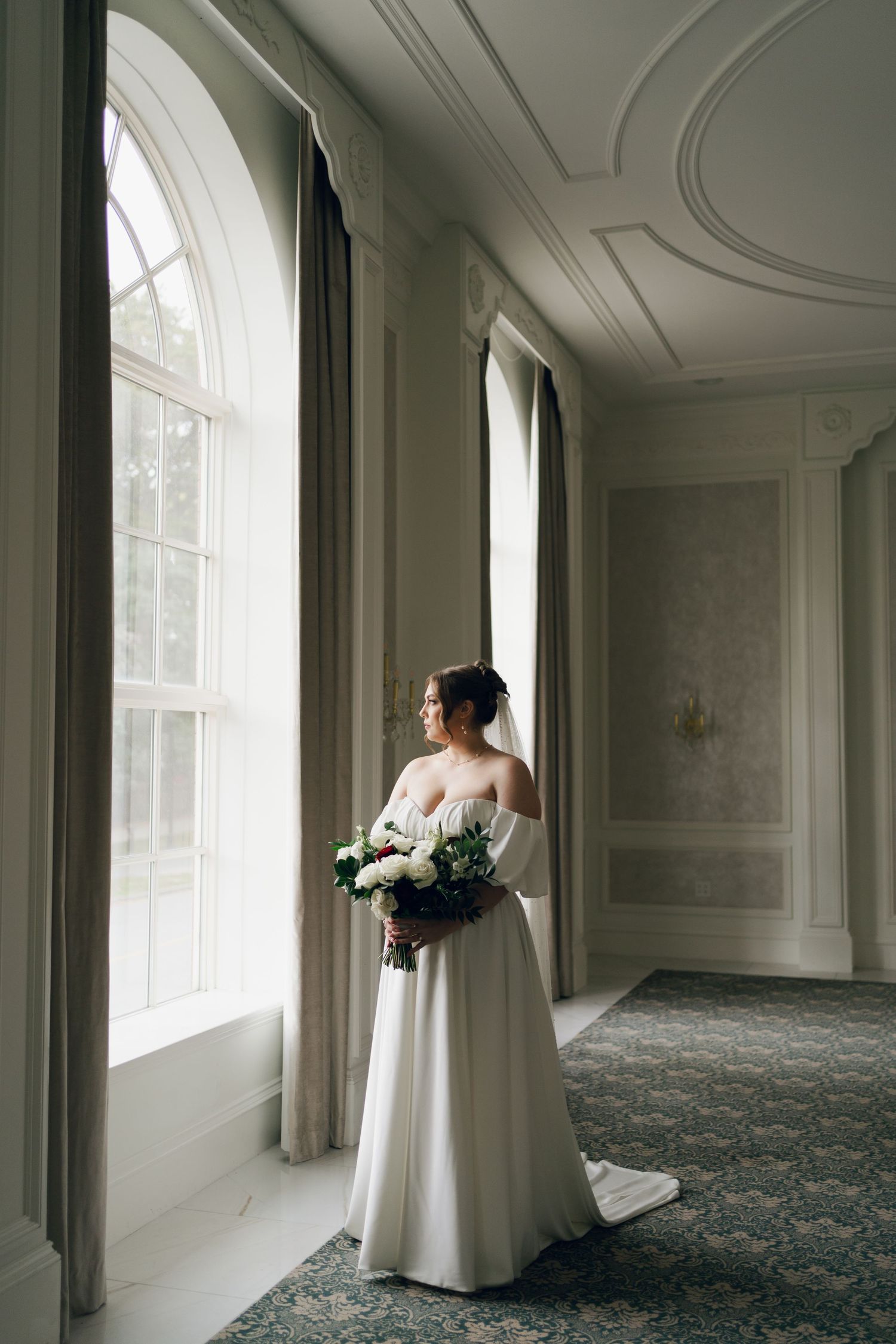 A bride in a white wedding dress stands near large windows in an elegant room with ornate molding and patterned carpet at Lord Nelson Hotel.