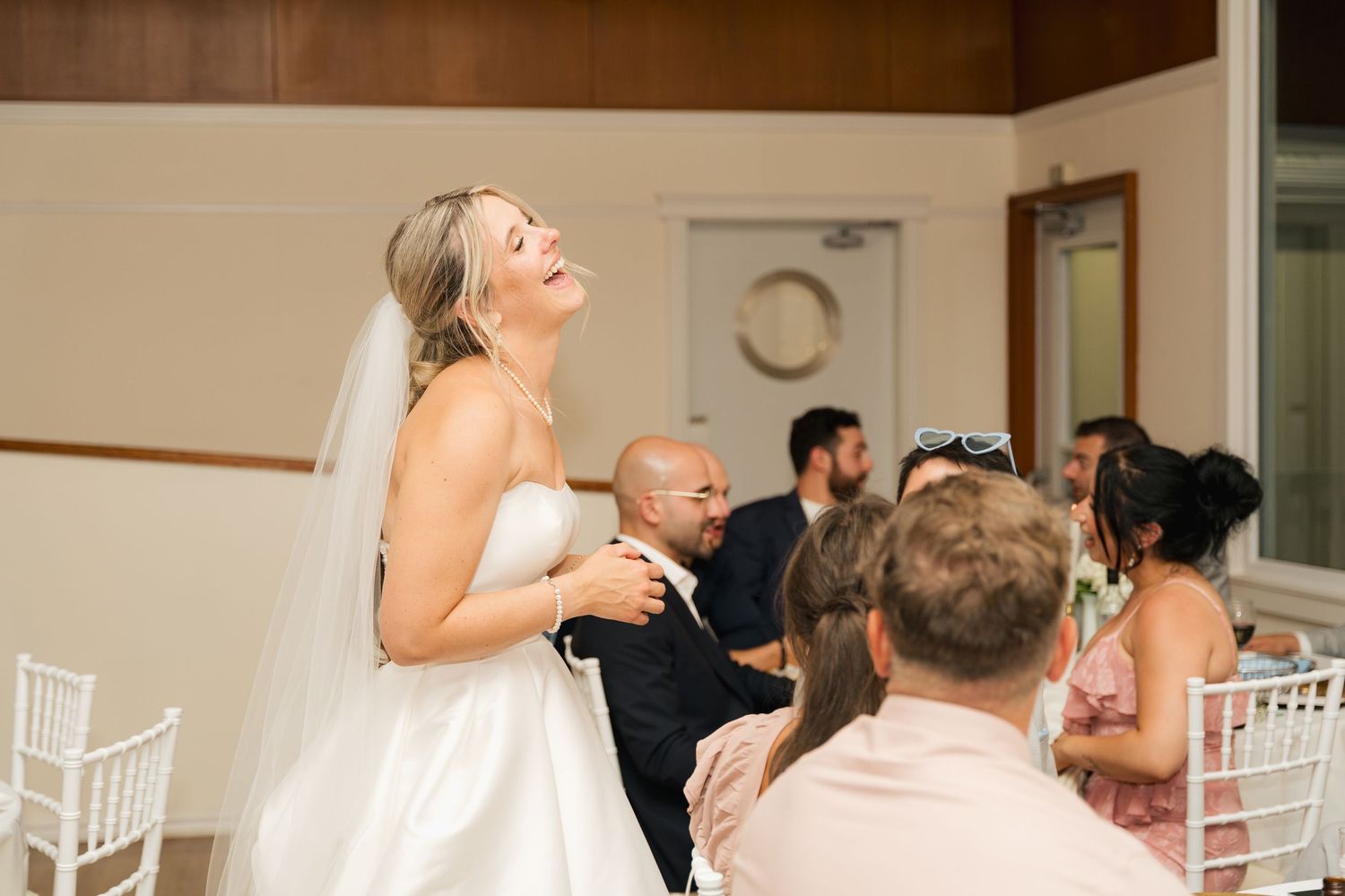 A bride laughs with joy during her wedding reception surrounded by guests in white chairs in Halifax, Nova Scotia.