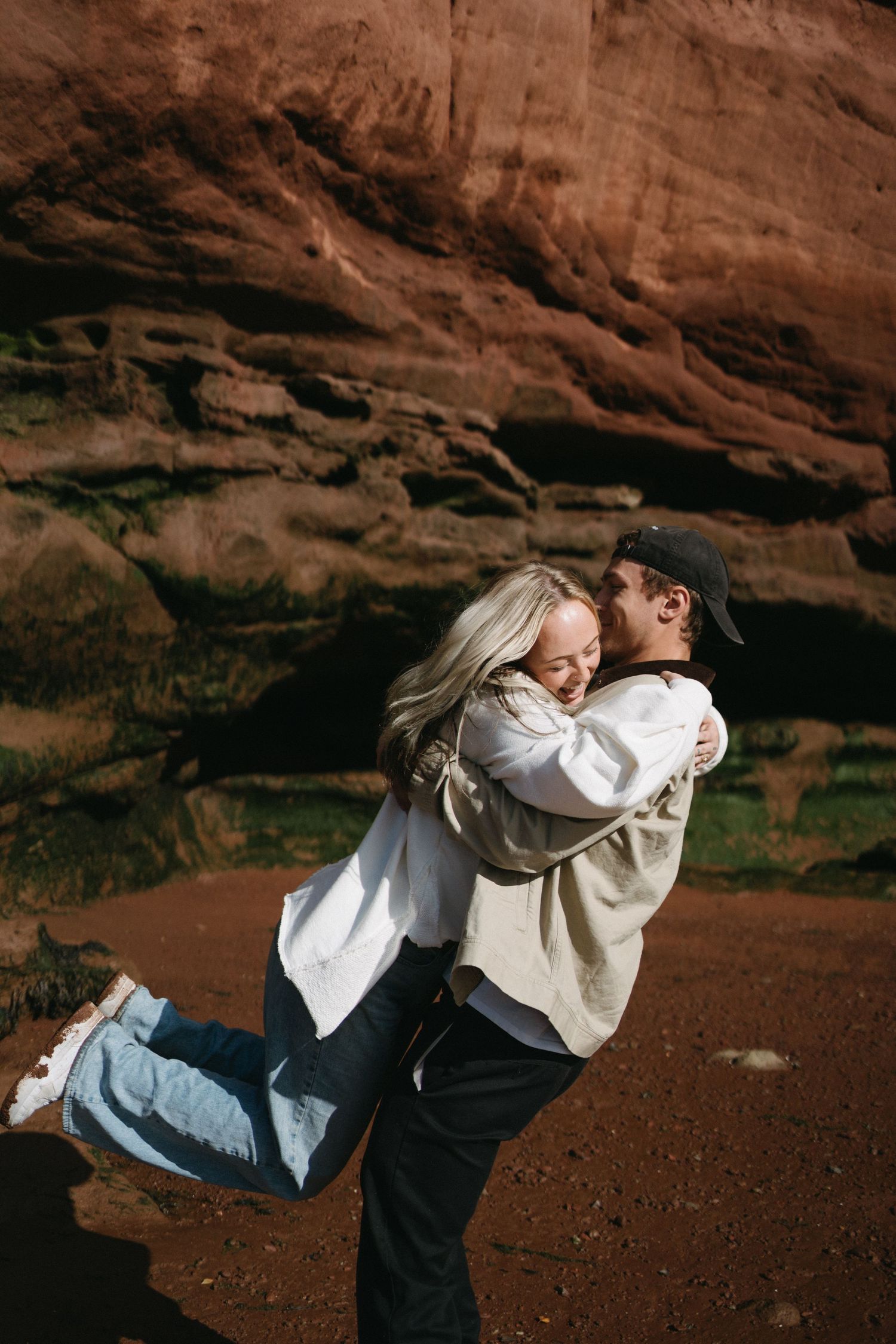 A couple embraces playfully against a backdrop of dramatic red rock formations in Nova Scotia.