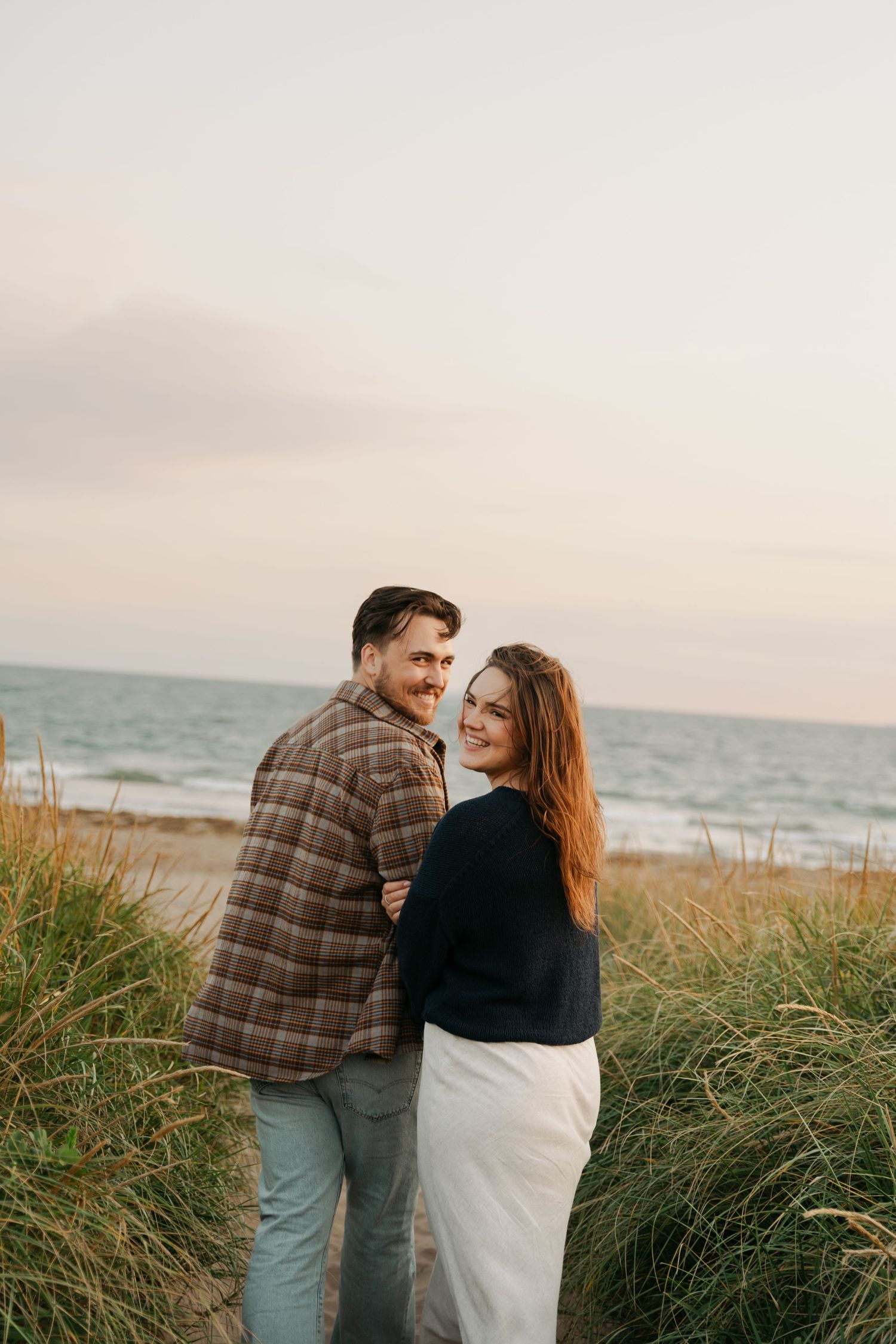 A couple embraces while walking along a coastal beach path with tall grass in Halifax, Nova Scotia and ocean views at sunset.