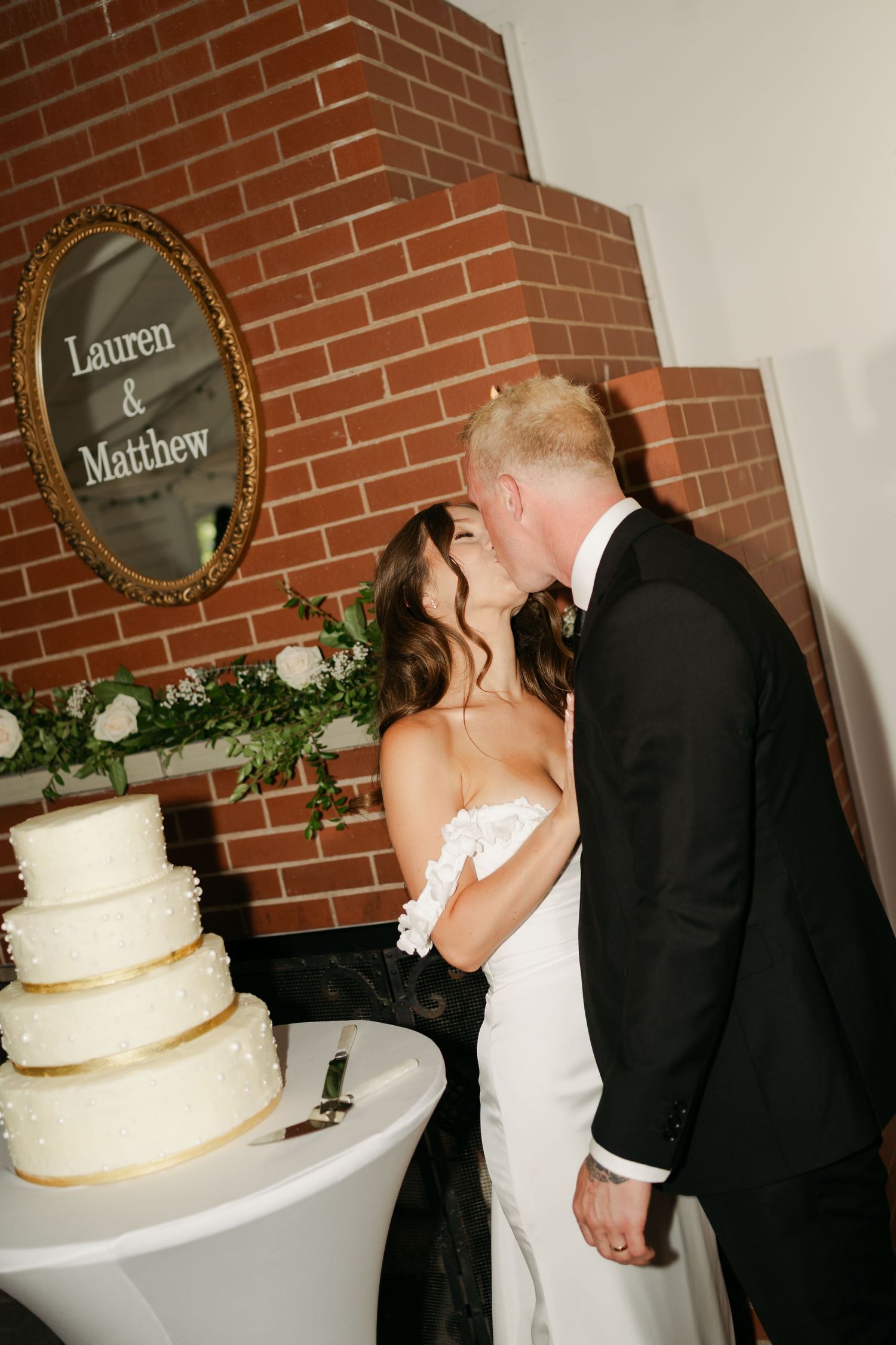 Wedding couple shares an intimate moment while cutting their white tiered cake next to a brick wall with decorative name plates in Halifax.