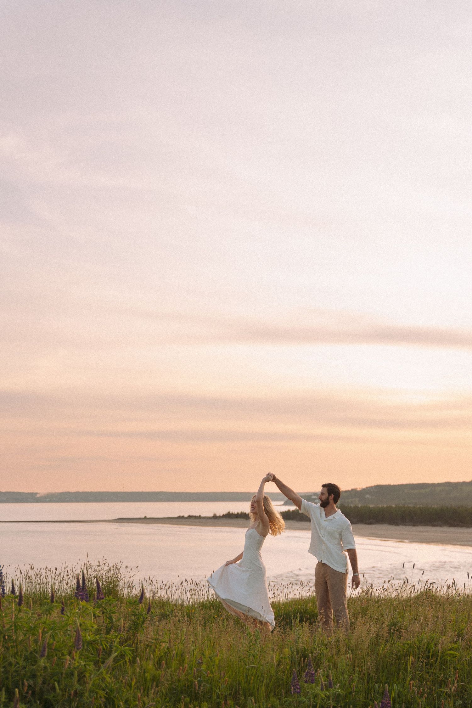 A couple dances together on a hilltop meadow with the ocean and sunset sky as their backdrop in Halifax, Nova Scotia.