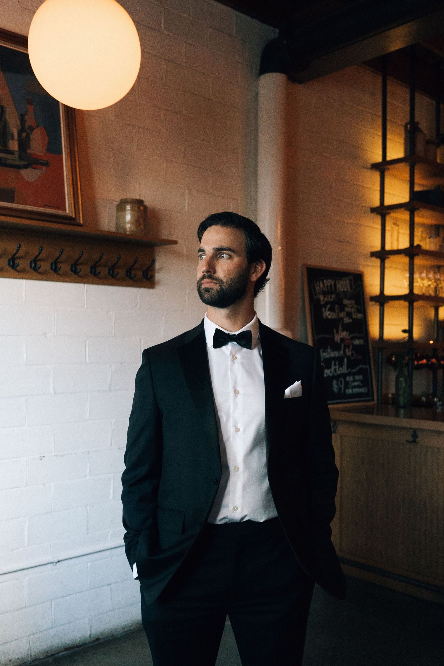 A groom in classic tuxedo poses against white brick wall with dramatic lighting in Halifax, Nova Scotia.