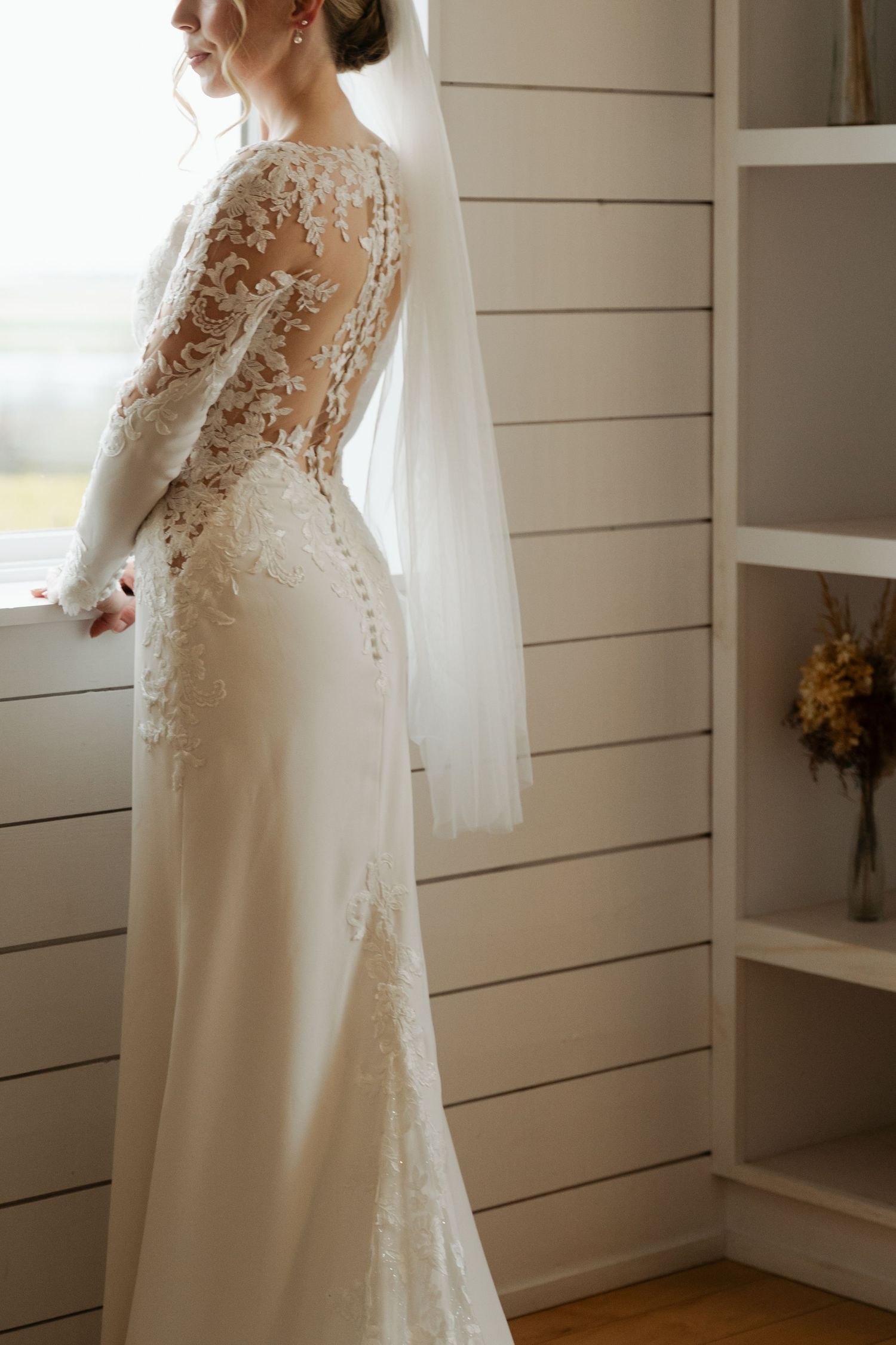 Elegant white wedding dress with delicate lace details and sheer back panel displayed against white shiplap wall at Lightfoot & Wolfville.