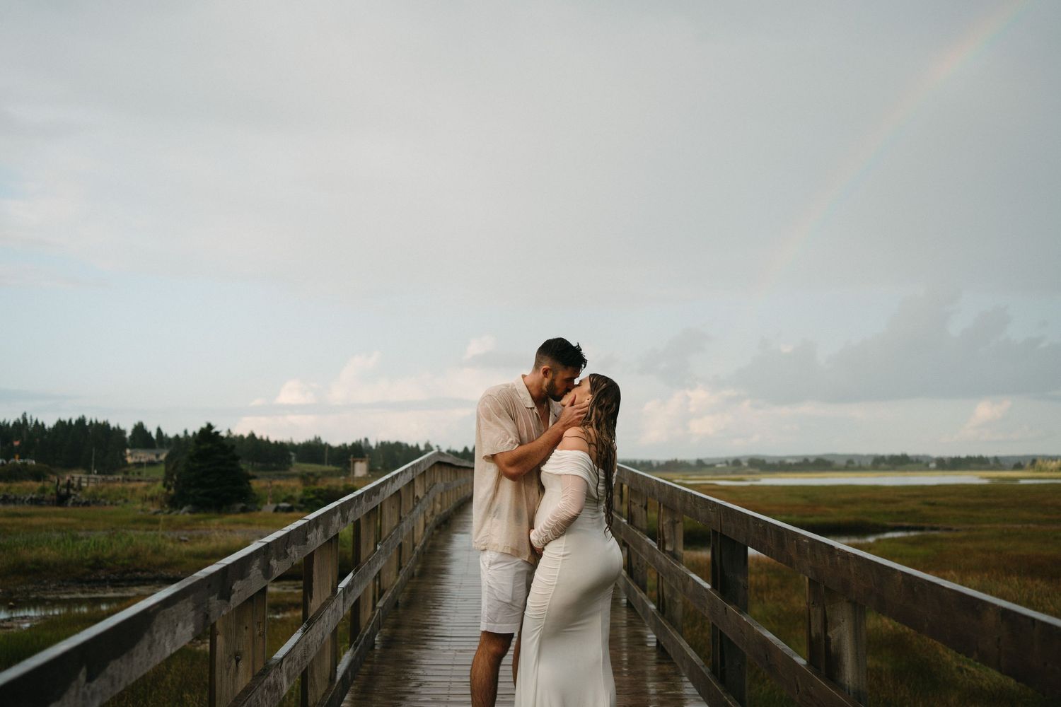 A couple shares a romantic moment on a wooden boardwalk overlooking a serene coastal marsh landscape in Halifax, Nova Scotia.