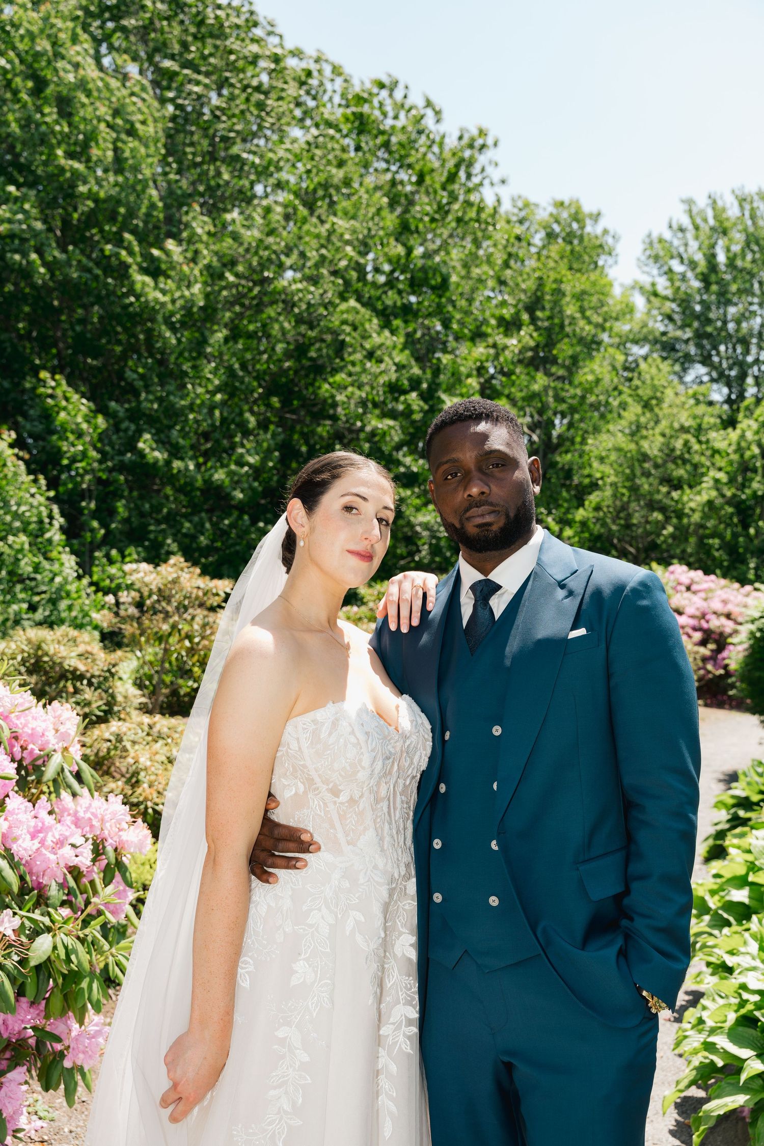 A couple poses together in a garden setting with pink flowers and lush greenery on their wedding day in Halifax, Nova Scotia.