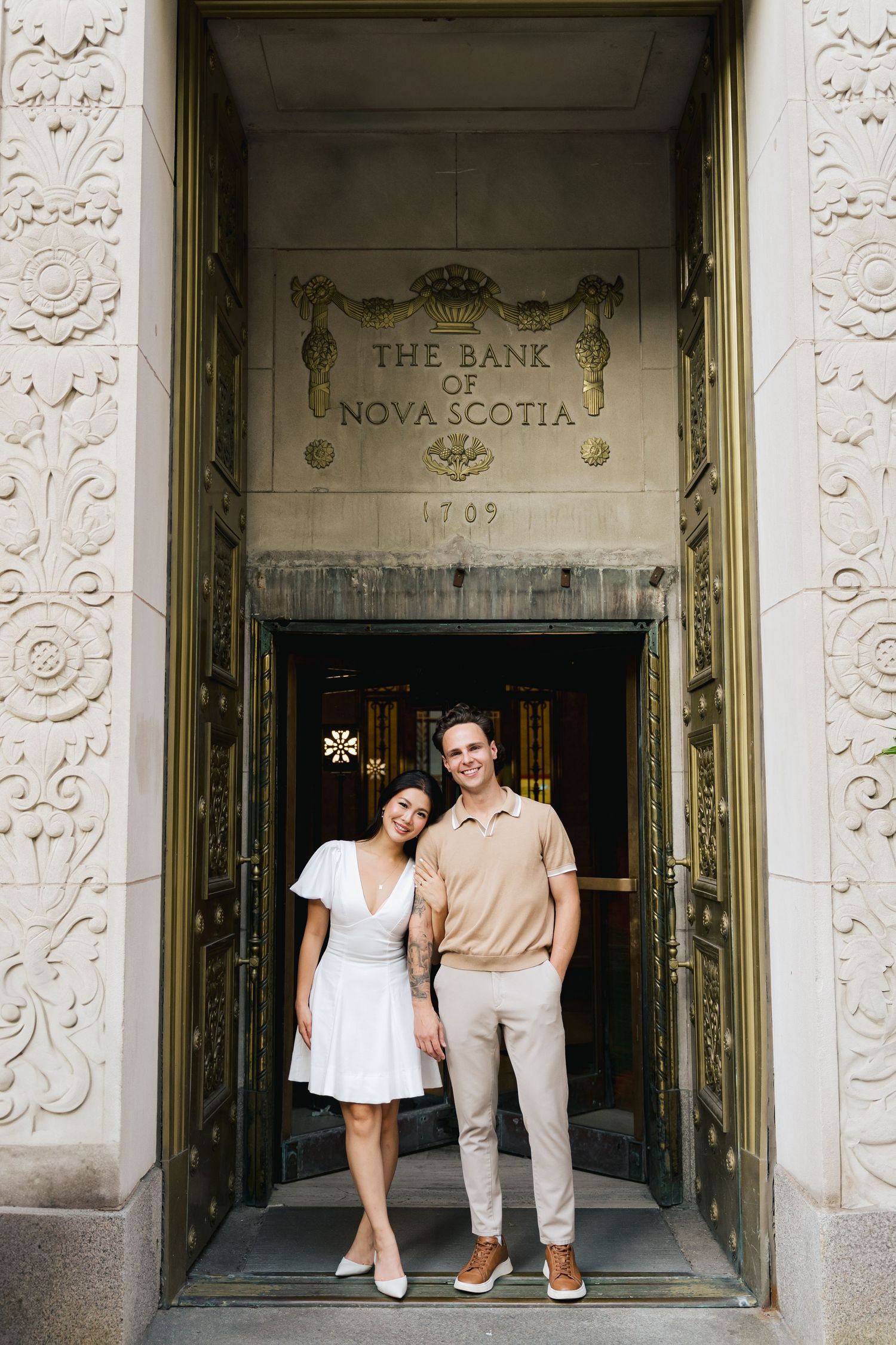A couple poses together in front of an ornate historic bank building entrance in downtown Halifax, Nova Scotia with decorative stone.