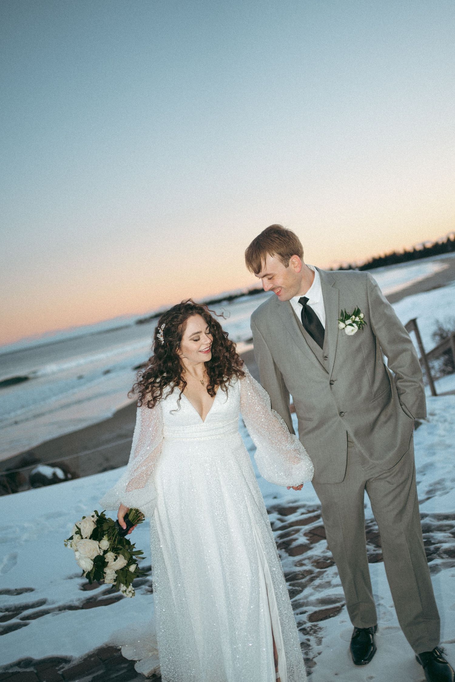 A couple in wedding attire walks along a snowy beach at sunset with white flowers and a soft winter glow at White Beach Resort, Nova Scotia.