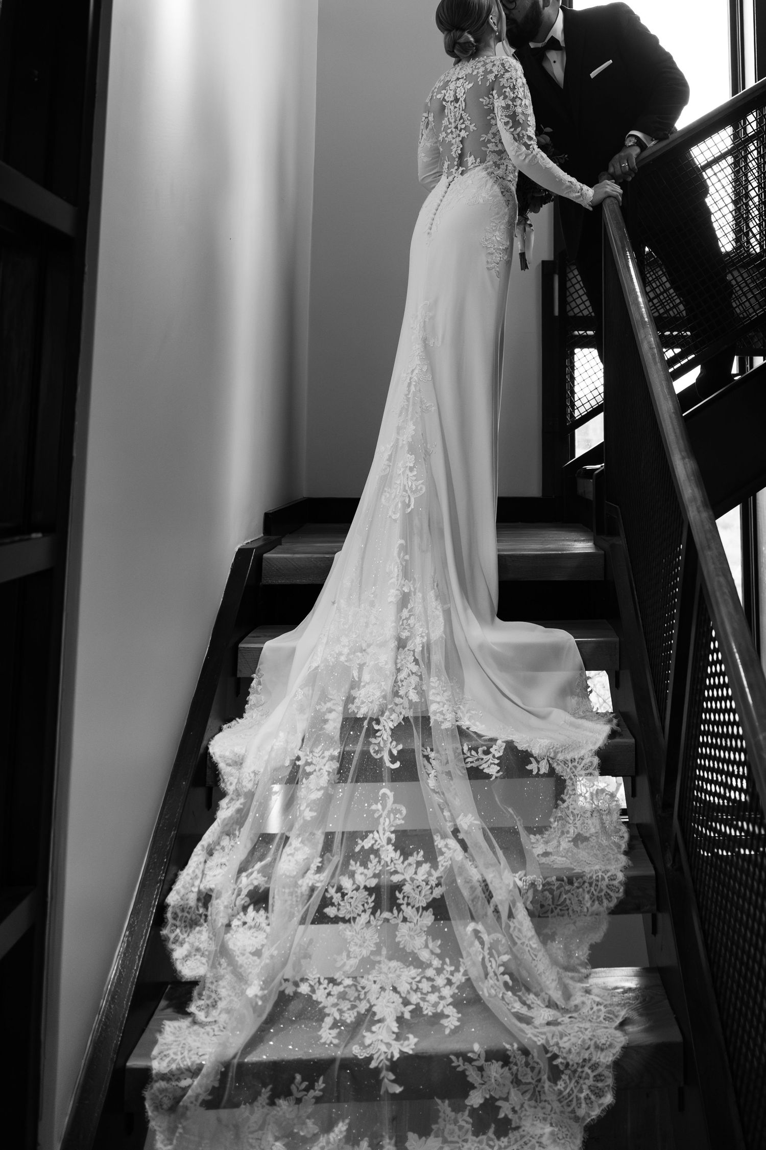 A wedding dress with an elegant lace train cascades down a dark staircase in a dramatic black and white photograph at Lightfoot & Wolfville.