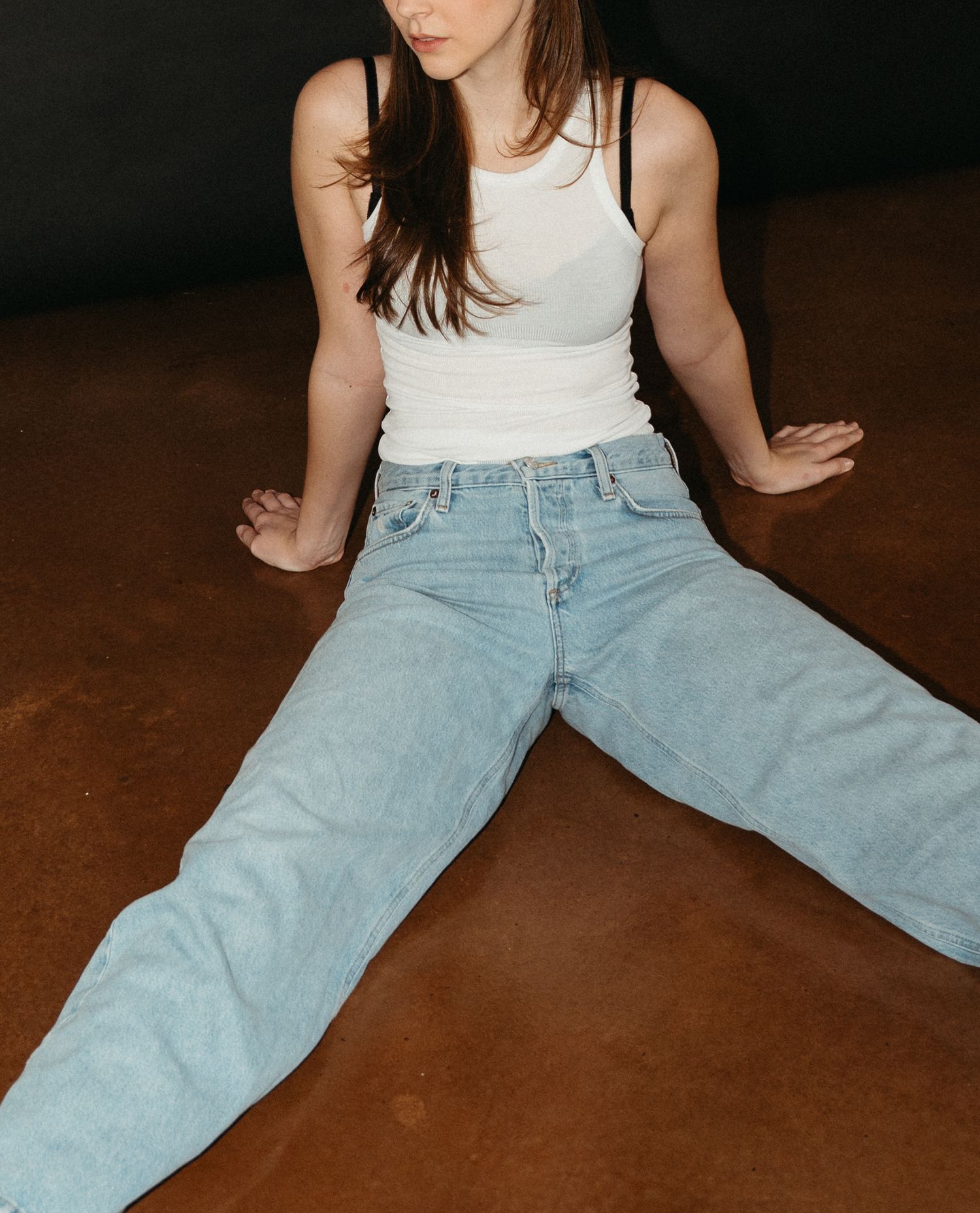 Person in light colored top and light wash jeans sits on dark floor in artistic fashion photography series in Halifax, Nova Scotia.