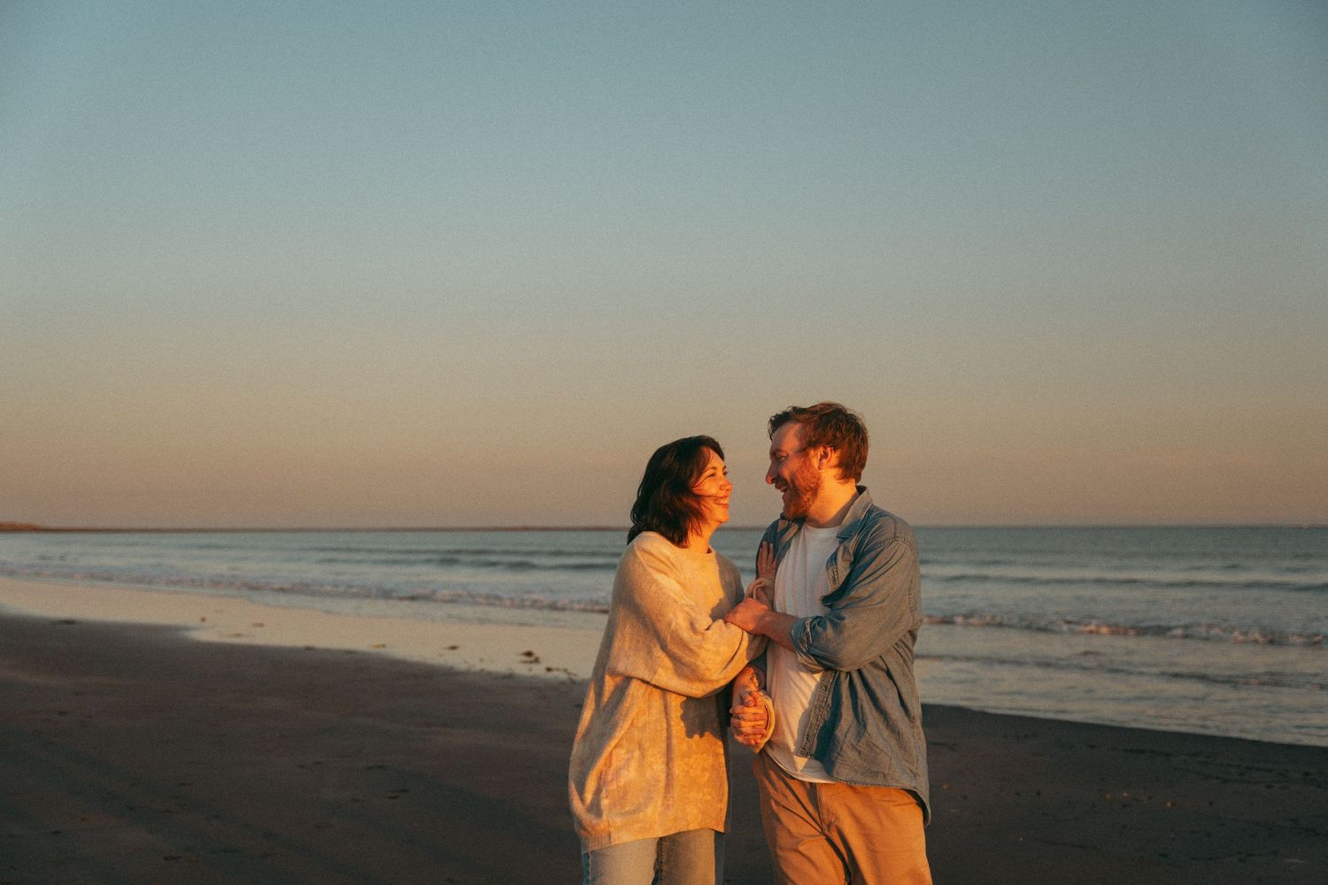 A couple shares a romantic moment at sunset on a peaceful beach in Halifax, Nova Scotia with golden light illuminating the scene.