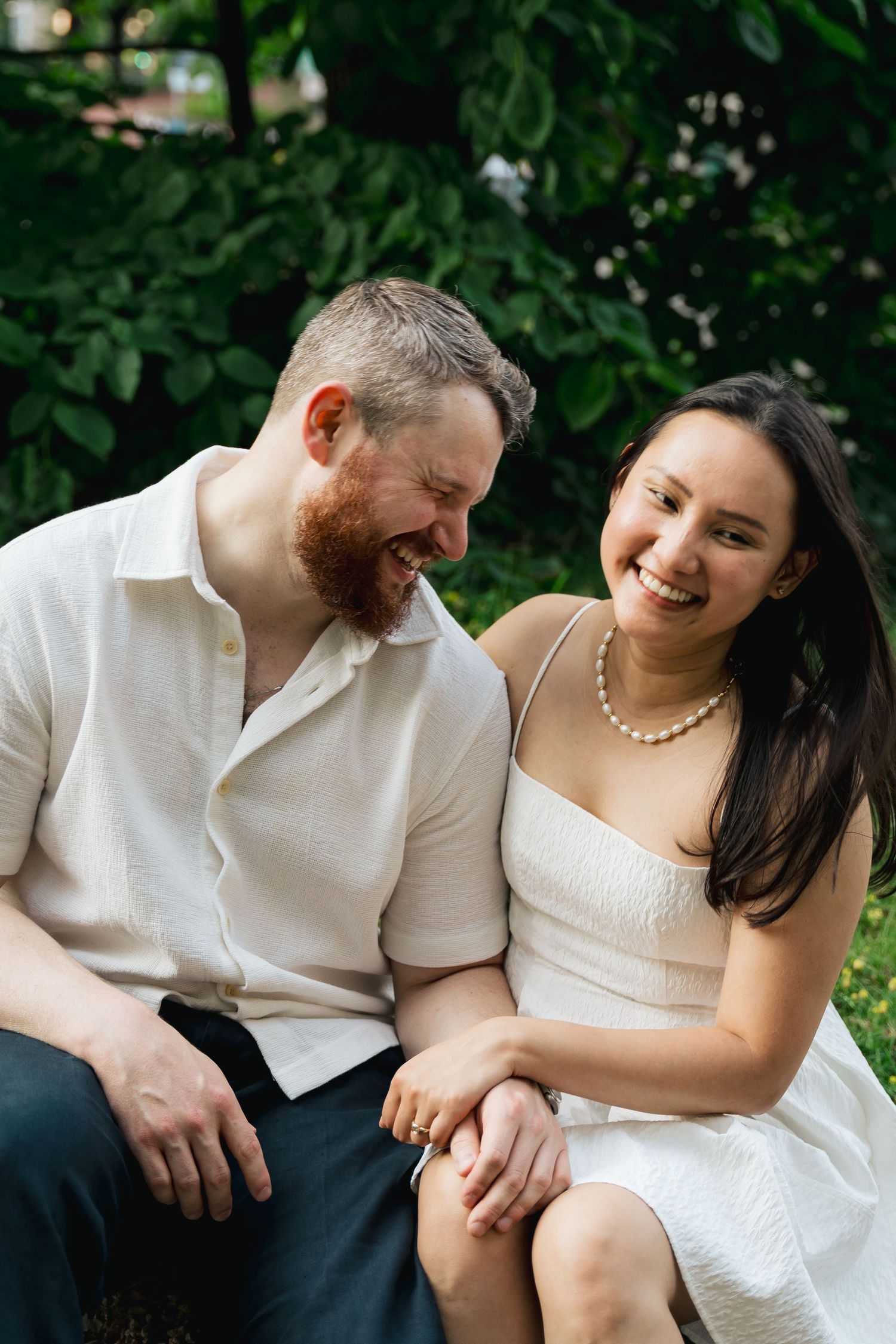 A couple in white clothing shares joyful laughter while sitting together outdoors in a natural setting in Halifax, Nova Scotia.