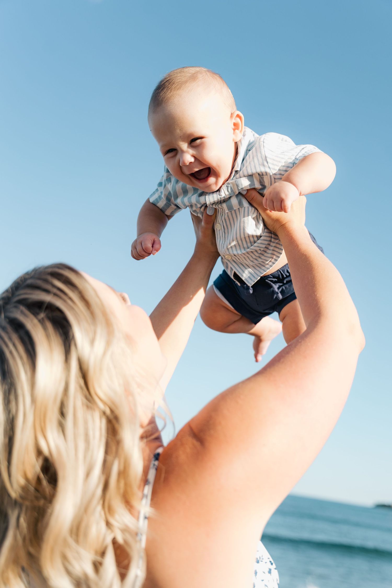 A parent lifts a joyful baby wearing stripes high in the air against a bright blue ocean backdrop in Halifax, Nova Scotia.