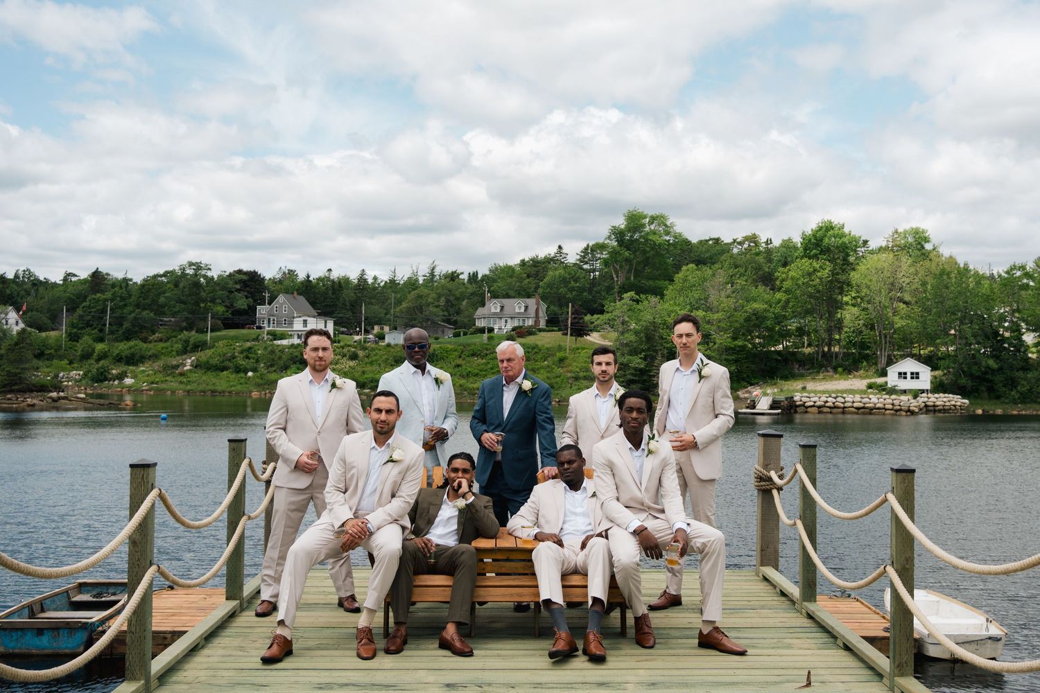 Wedding party members sit together on a wooden dock overlooking a serene lake on a summer day in Halifax, Nova Scotia.