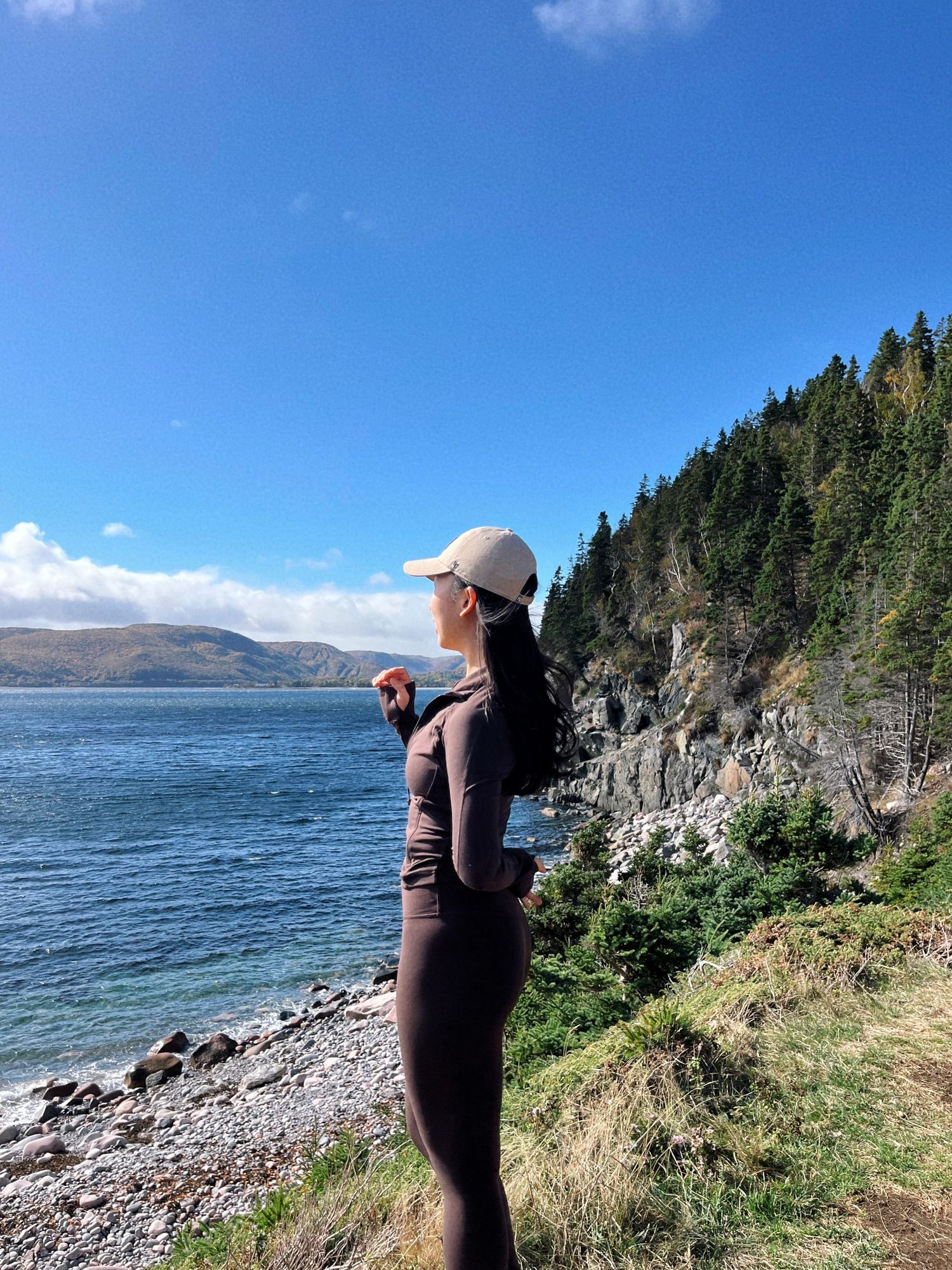 A rocky coastal landscape with evergreen trees and mountains overlooking a serene lake on a sunny day in Cape Breton, Nova Scotia.