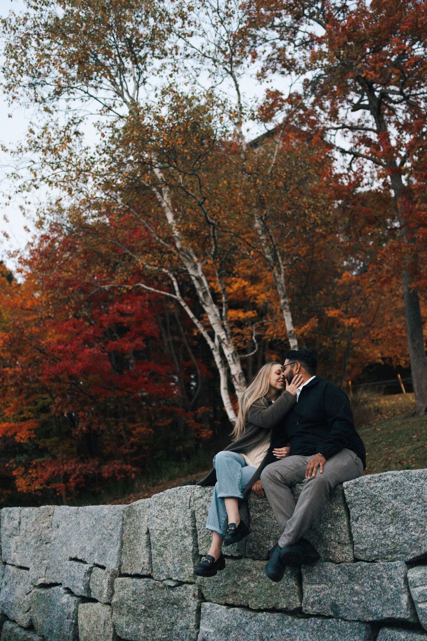 Two people sit together on a stone wall surrounded by vibrant autumn foliage in a park setting in Halifax, Nova Scotia.